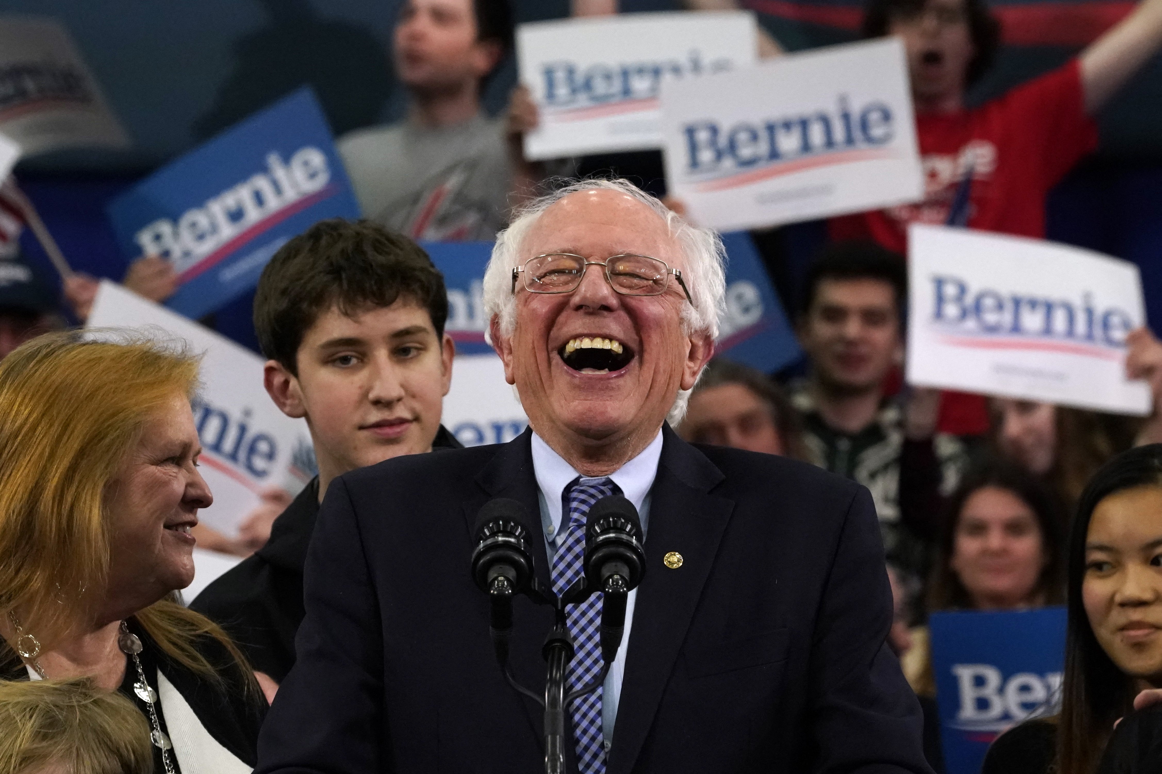 Smilende mann i dress på et rally, omgitt av folkemengder som holder «Bernie»; tegn