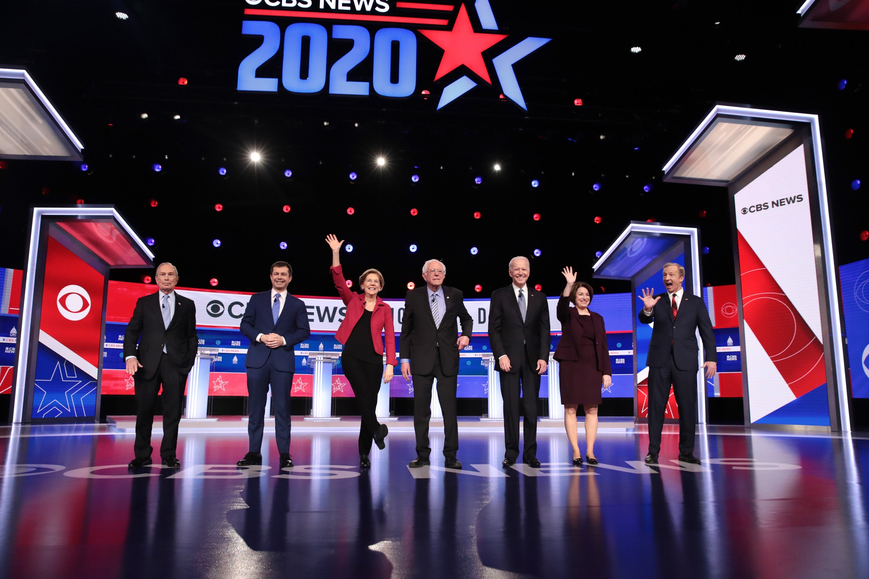 Gruppe individer i forretningsantrekk på en debattscene, vinker til et publikum under CBS News 2020-skilting