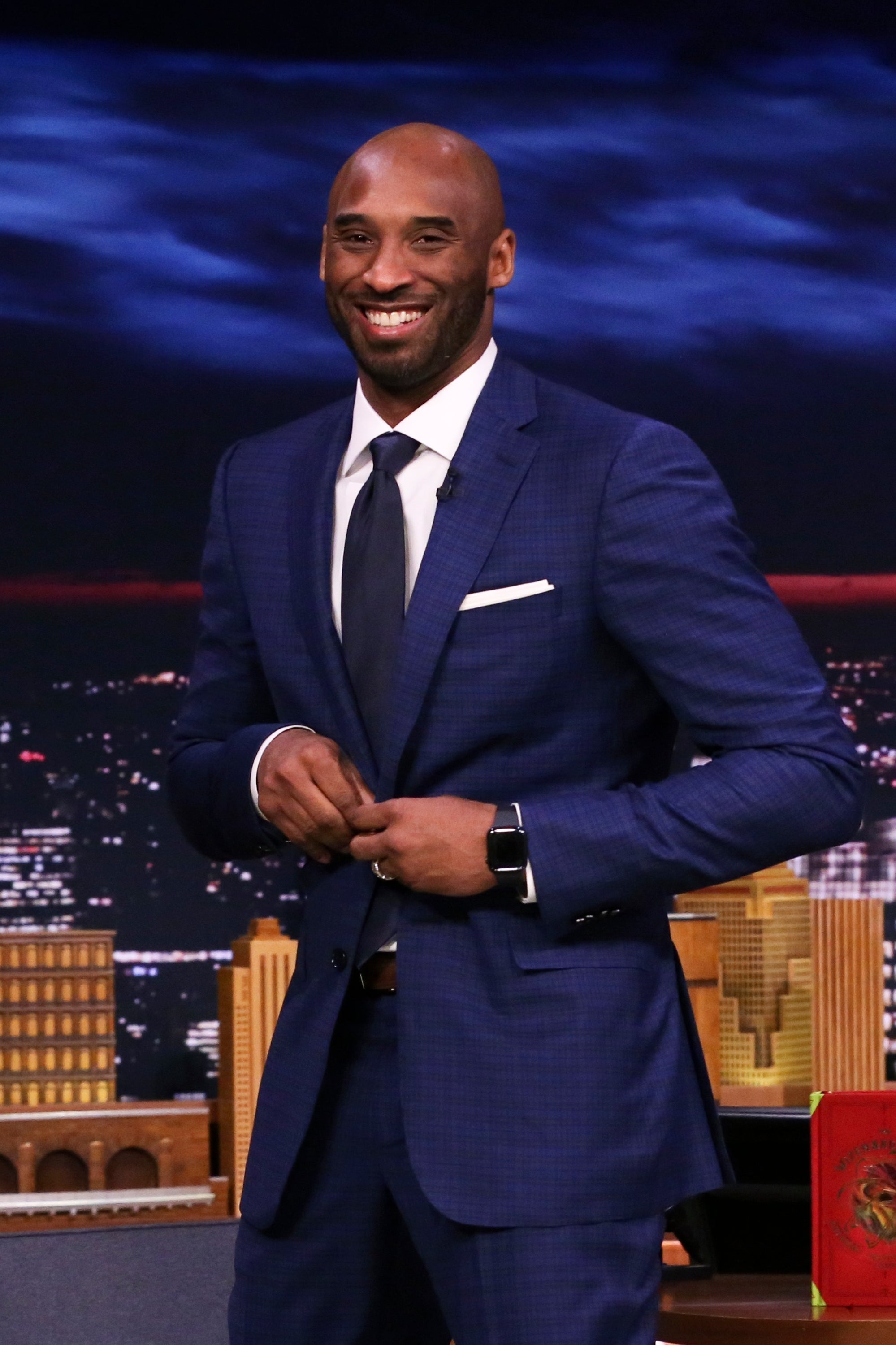 Mann i skarp dress smiler på et talkshowsett med bybilde i bakgrunnen