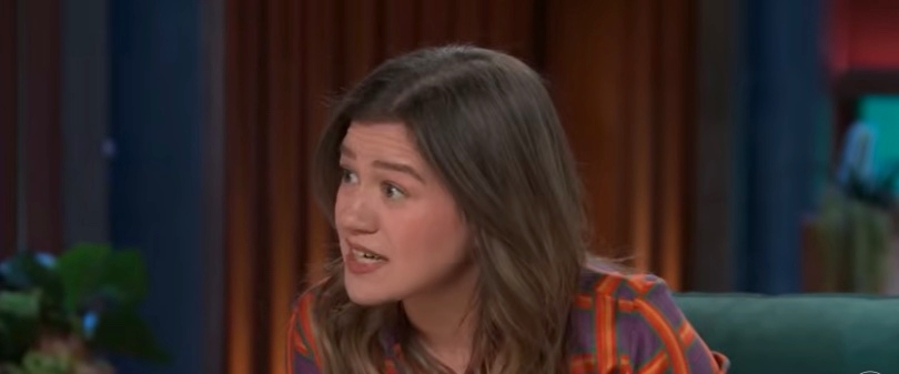 Person with long hair gestures while speaking on a talk show set