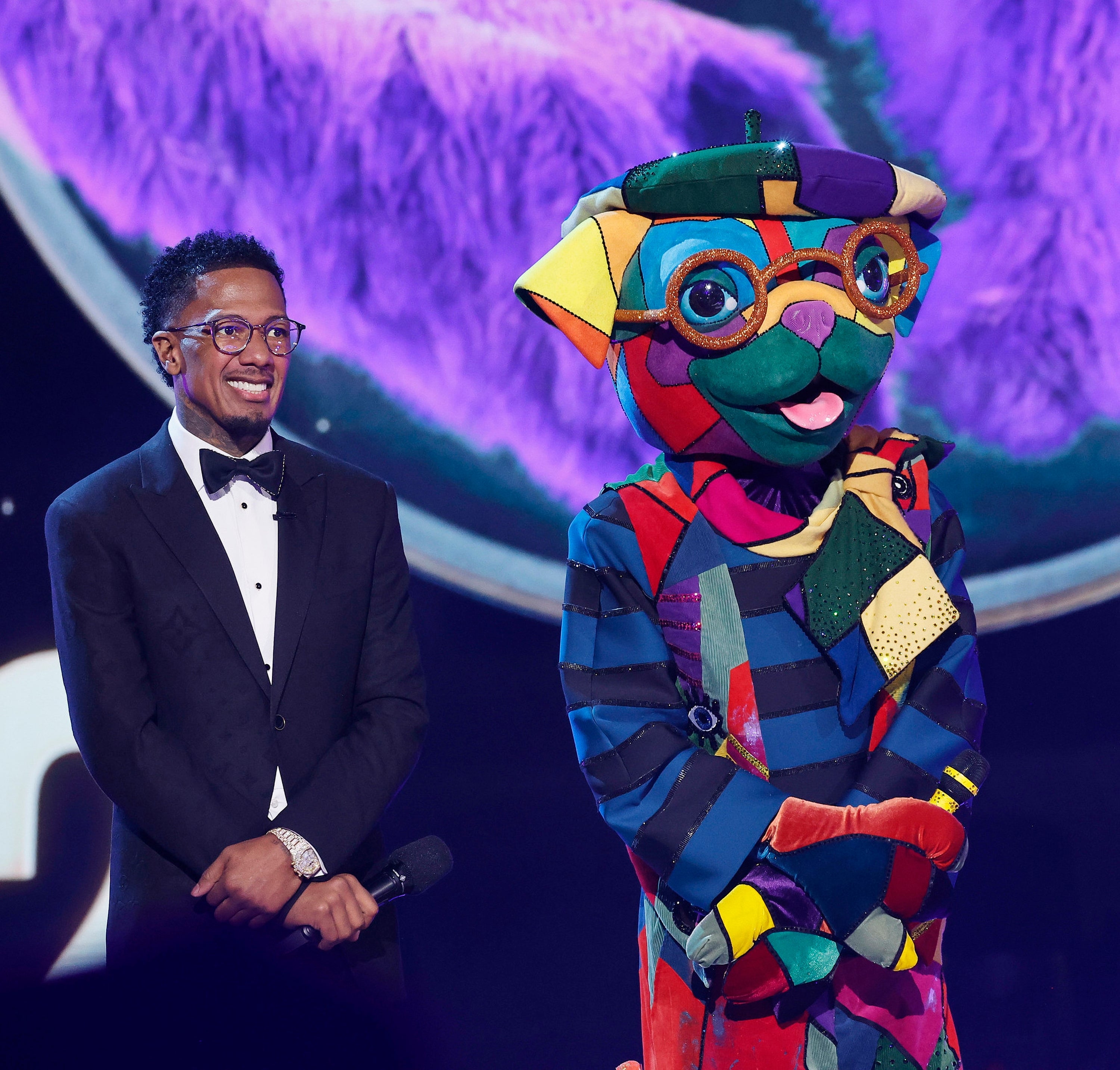 Vert i smoking med briller står ved siden av en fargerik lappeteppekostyme på scenen, muligens på et TV-arrangement