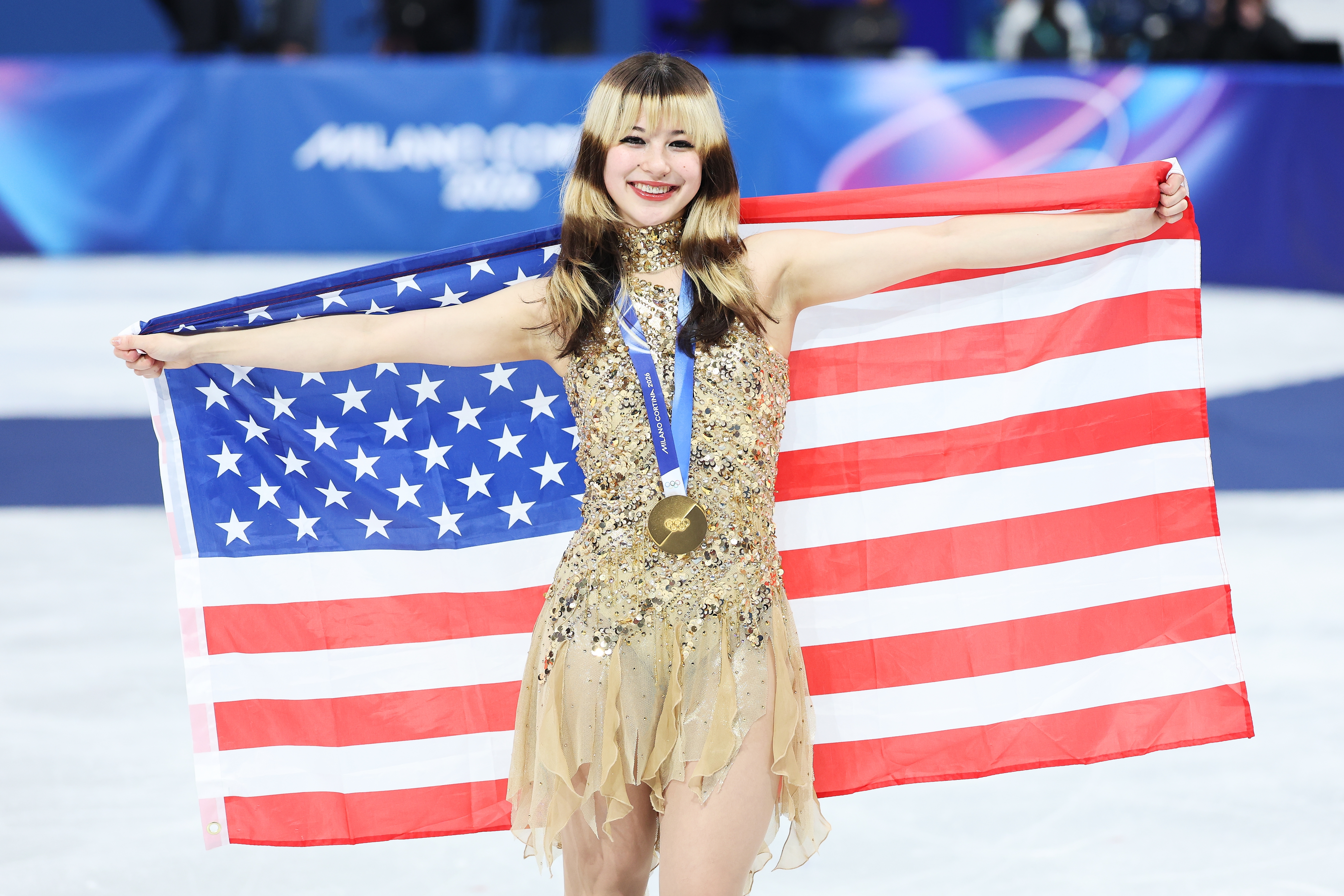 Figure skater successful  a golden  sequined outfit poses connected  ice, holding a U.S. emblem  and wearing a medal