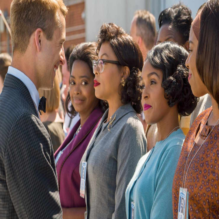 A group of women in 1960s office attire, with name badges, converse with a man in a suit, possibly a scene from a historical film or TV show