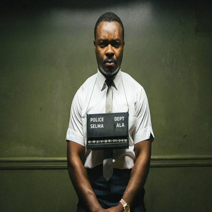 A man in a 1960s police mugshot setting, wearing a dress shirt and tie, with "Selma" and "Dept Ala" sign around his neck, depicting a historical scene