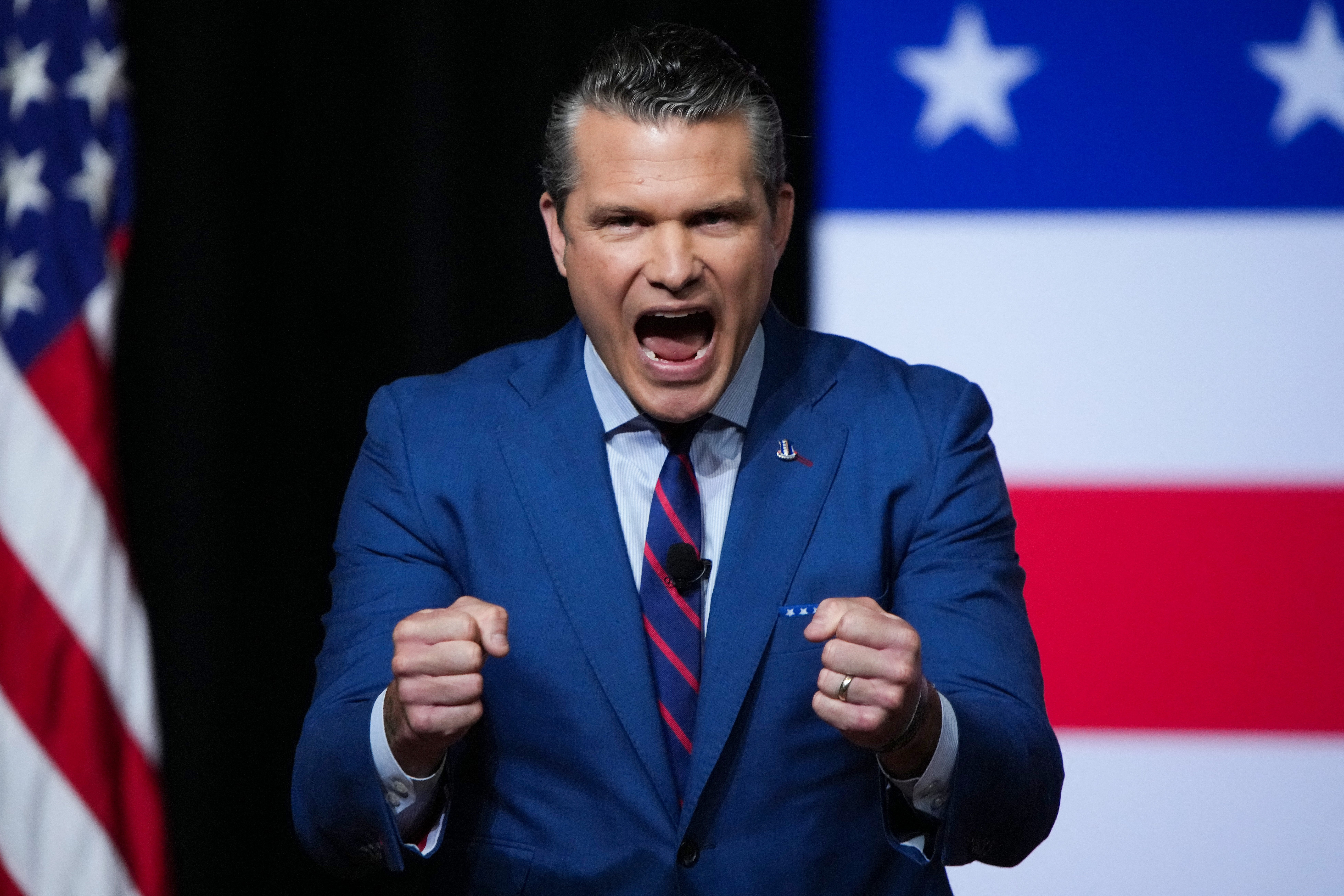 Pete Hegseth foran et amerikansk flaggbakgrunn, med munnen åpen og hendene utformet som to knyttnever holdt opp omtrent midt på brystet