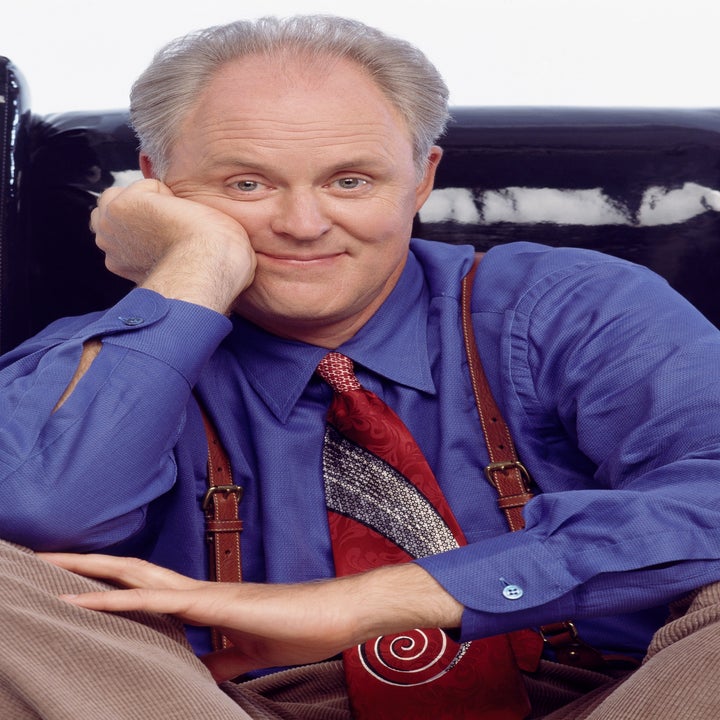 Pessoa sentada em uma cadeira, vestindo camisa azul e gravata estampada com suspensórios, sorrindo com o queixo apoiado na mão