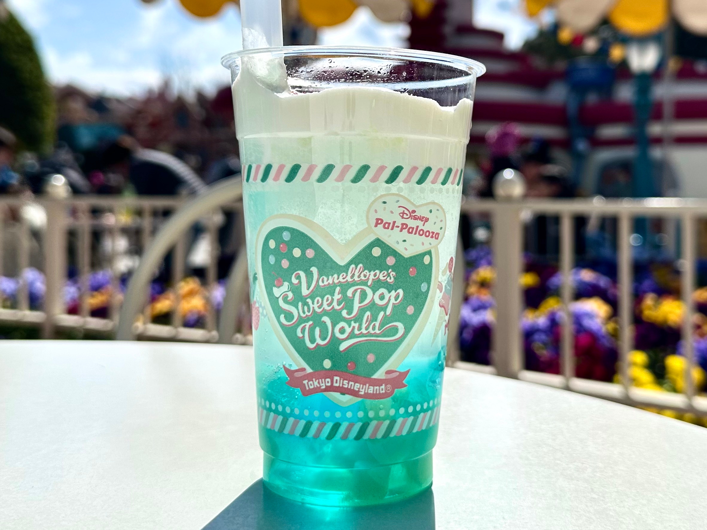 ディズニーランドで撮影された、ヴァネロペのスウィートポップワールドのソーダが入ったカップ。背景に遊園地の風景。