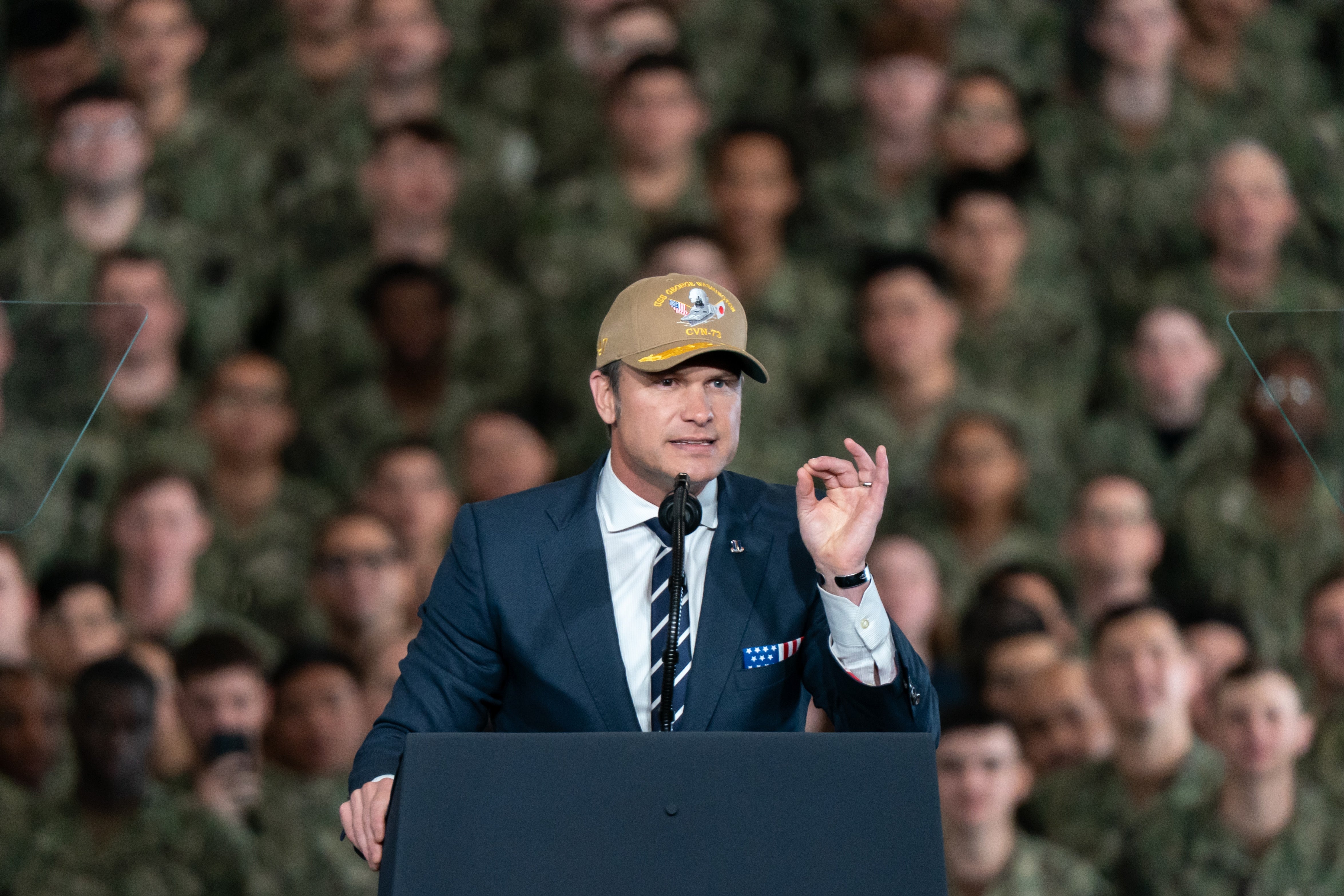 Pete Hegseth i dress og stripet slips, iført baseballcaps, snakker på et podium foran militært personell
