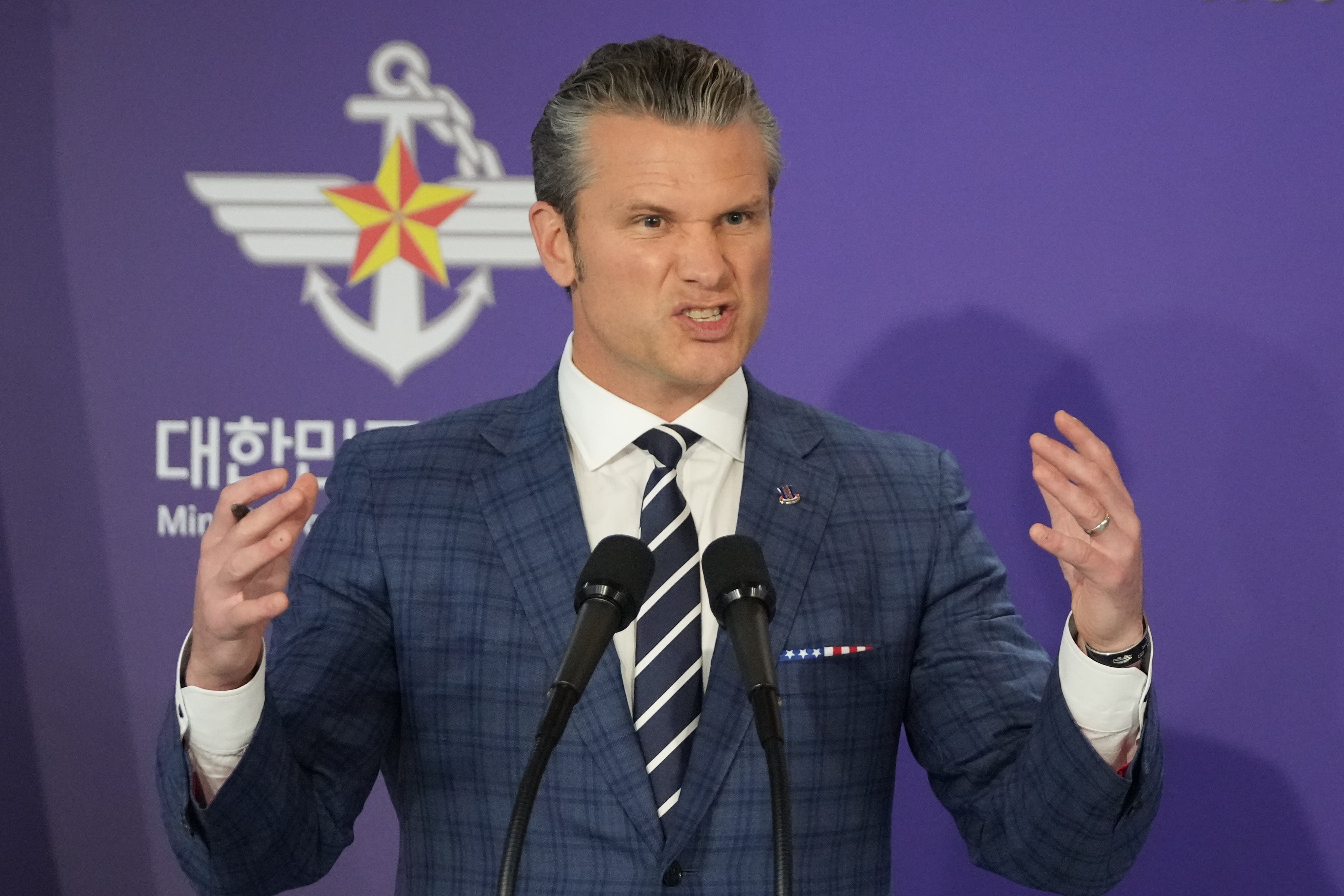 Pete Hegseth på et podium, gestikulerer med et anstrengt blikk mens han holder en tale, iført rutete dress, stripet slips og hvit skjorte. Militært emblem i bakgrunnen