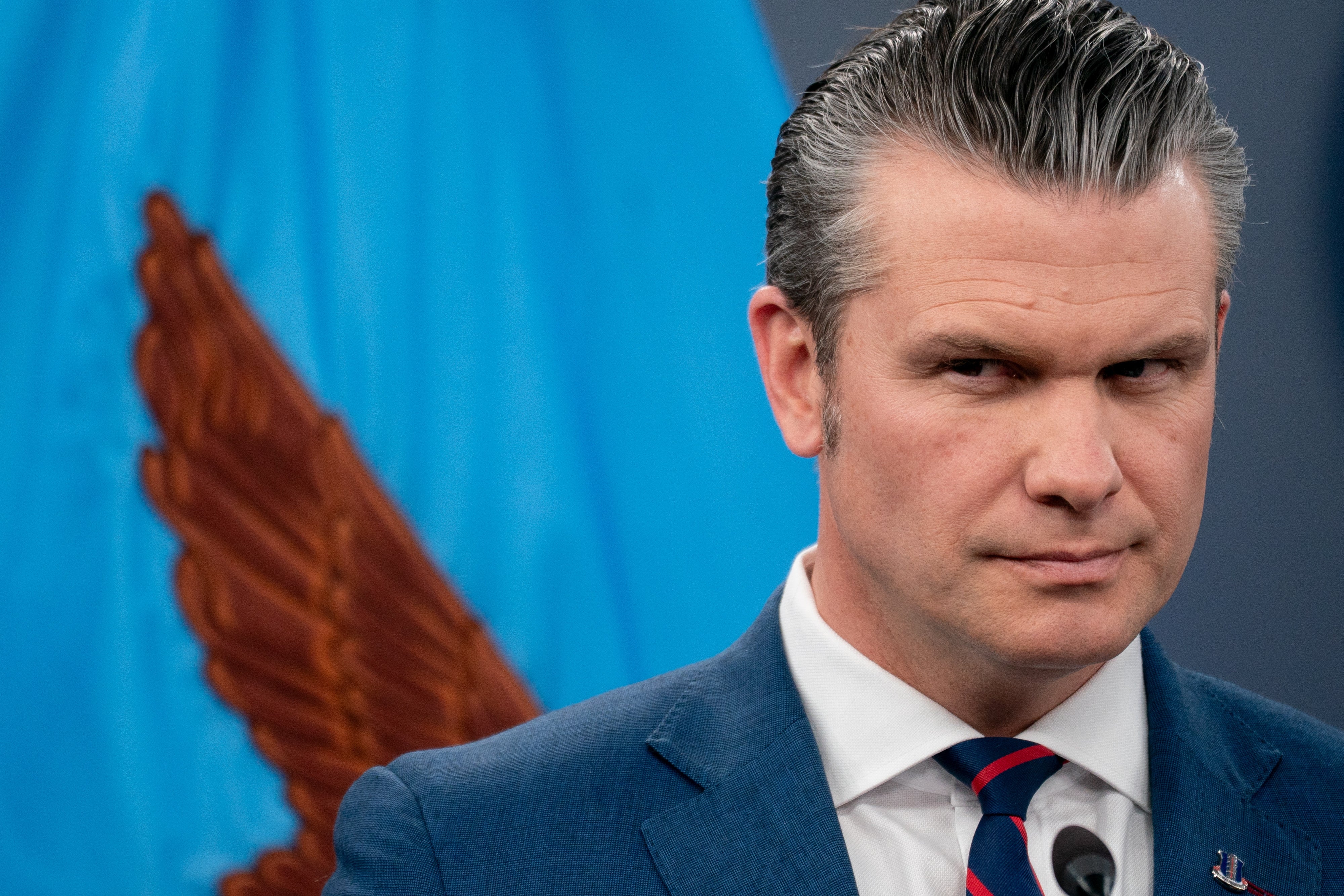 Pete Hegseth i dress står med et alvorlig uttrykk, foran et ørneemblem. Vingen til ørnen på flagget ser ut som den kommer ut av skulderen hans