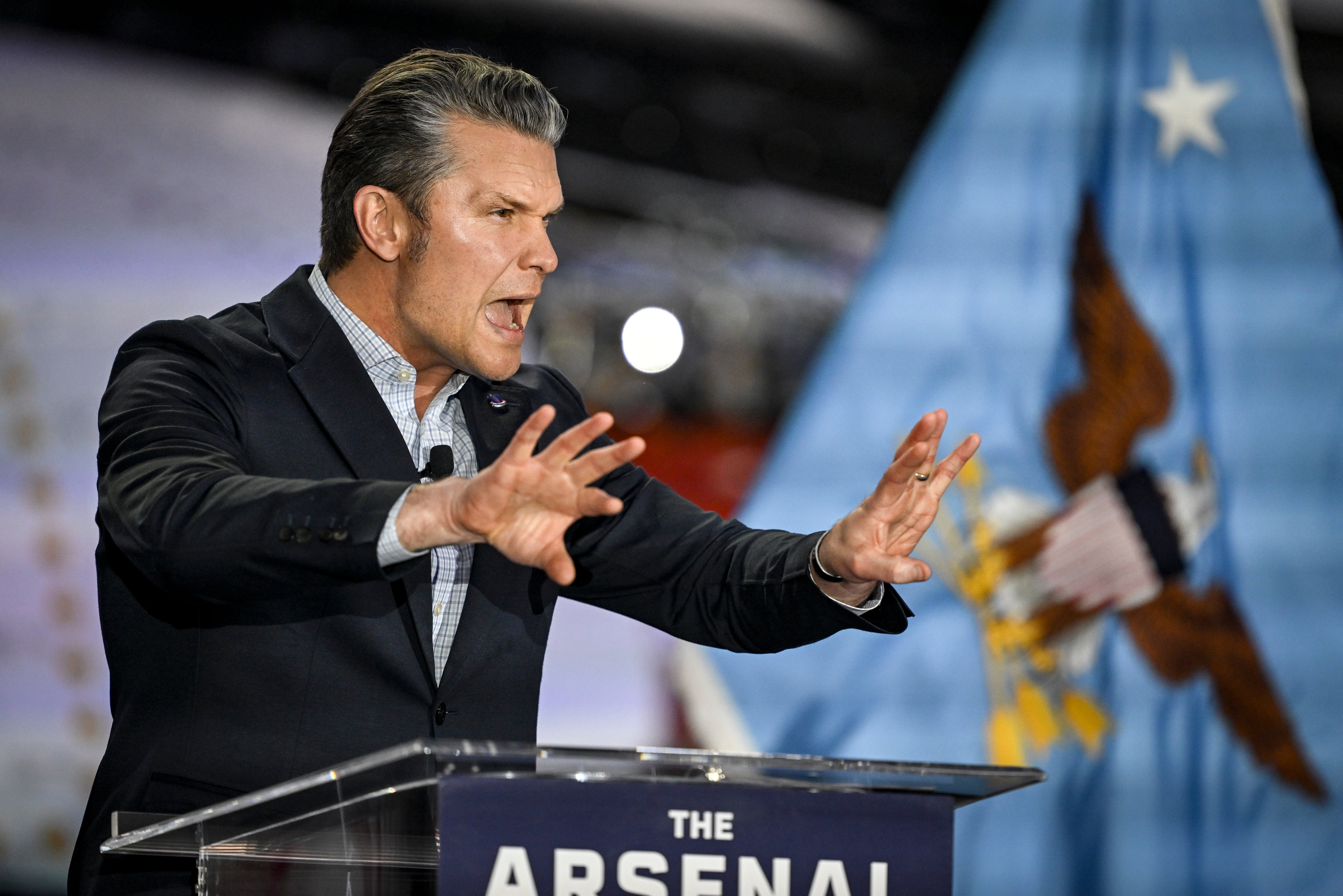 Pete Hegseth taler lidenskapelig ved et podium merket «ARSENALET»; gestikulerer med begge hender. Et ørn- og stjerneemblem er synlig i bakgrunnen. Det er en hvit, rund refleksjon i bildet rett foran ham