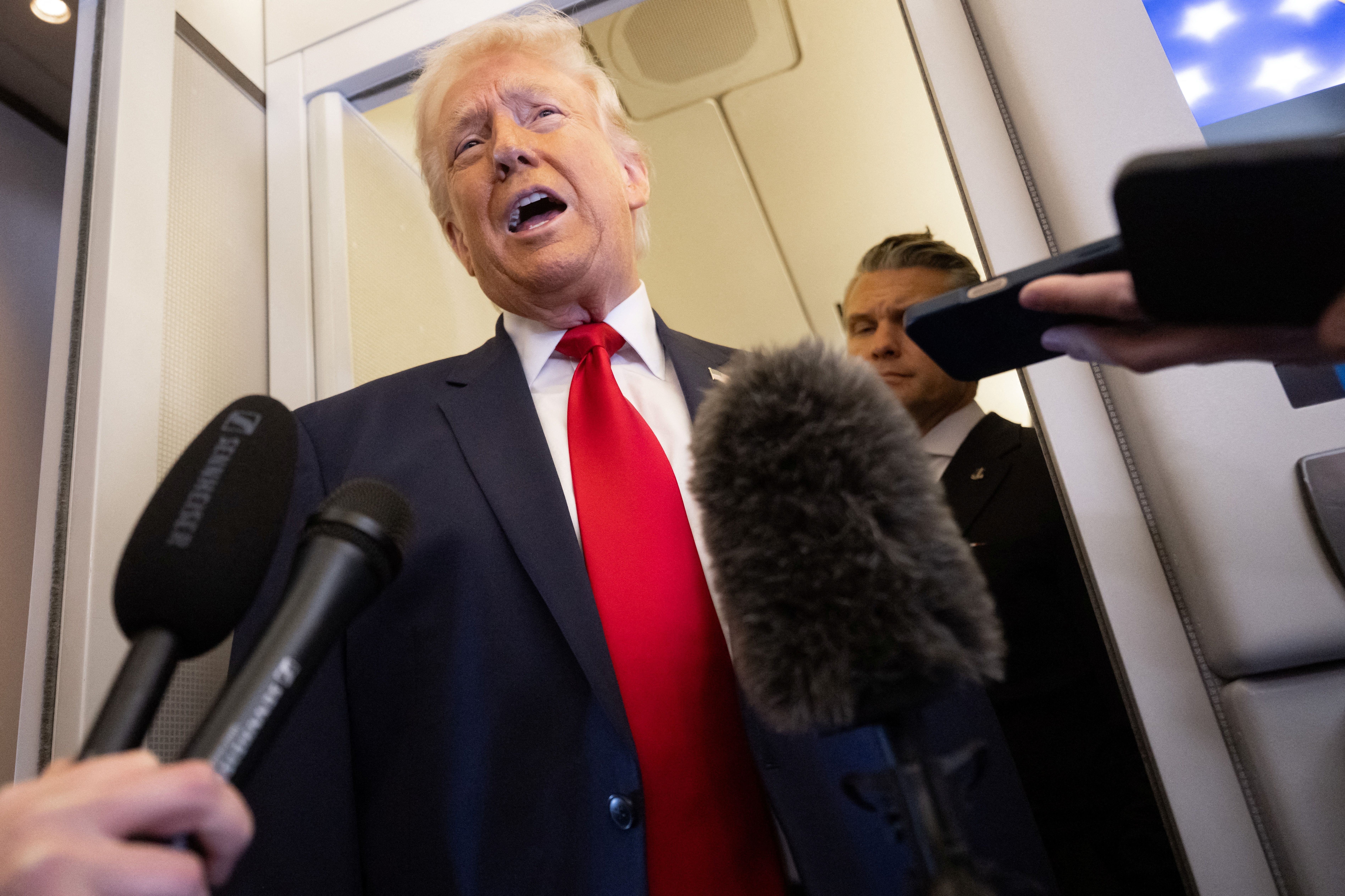 Donald Trump snakker til journalister, omgitt av mikrofoner, på et fly. Kledd i dress med rødt slips. Pete Hegseth blir presset inn i en døråpning bak seg med lukkede øyne