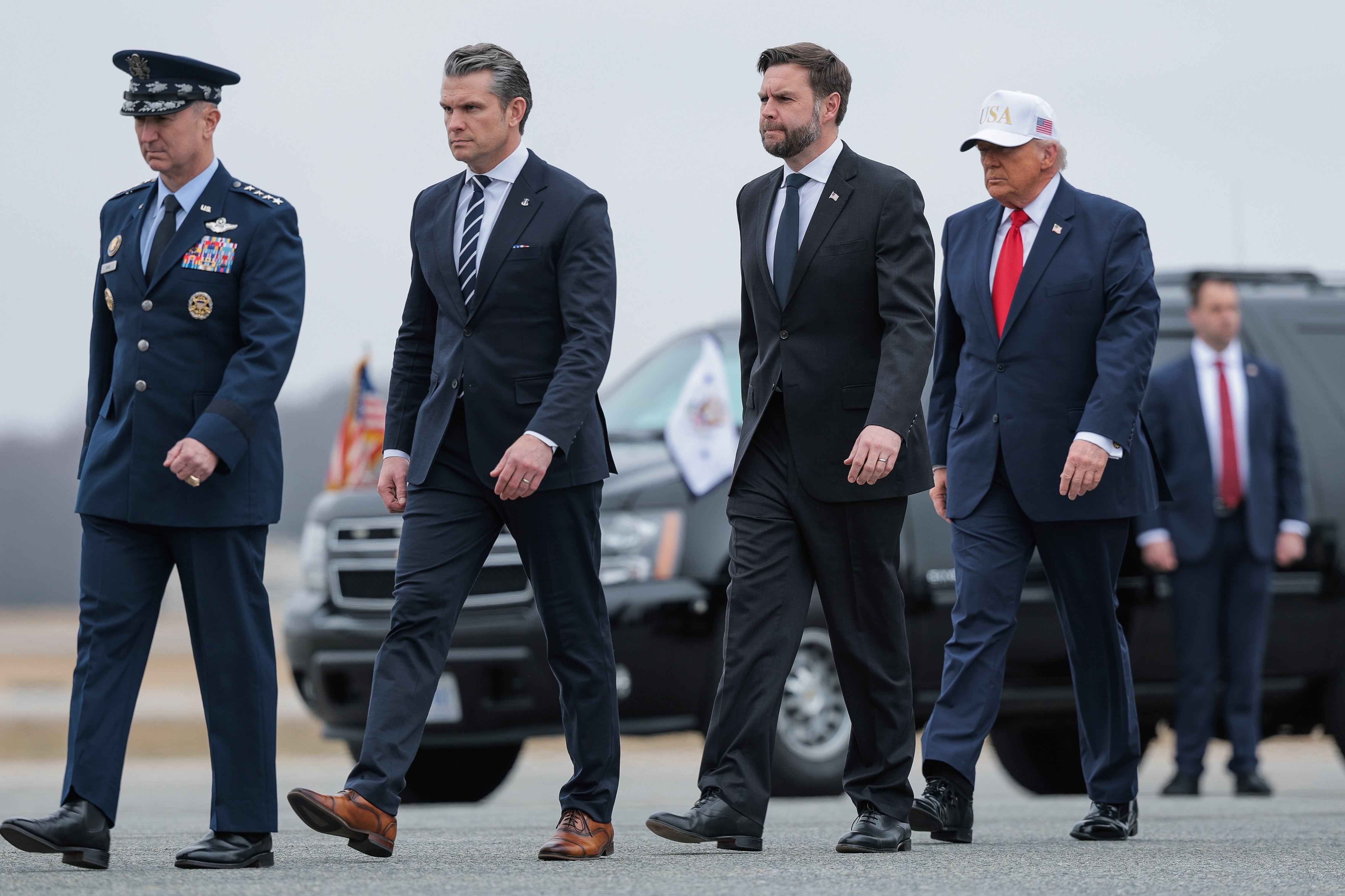 Et militærmedlem leder en prosesjon av Pete Hegseth, JD Vance og Donald Trump. De går i kø foran en svart SUV; Pete har på seg en svart dress og brune sko
