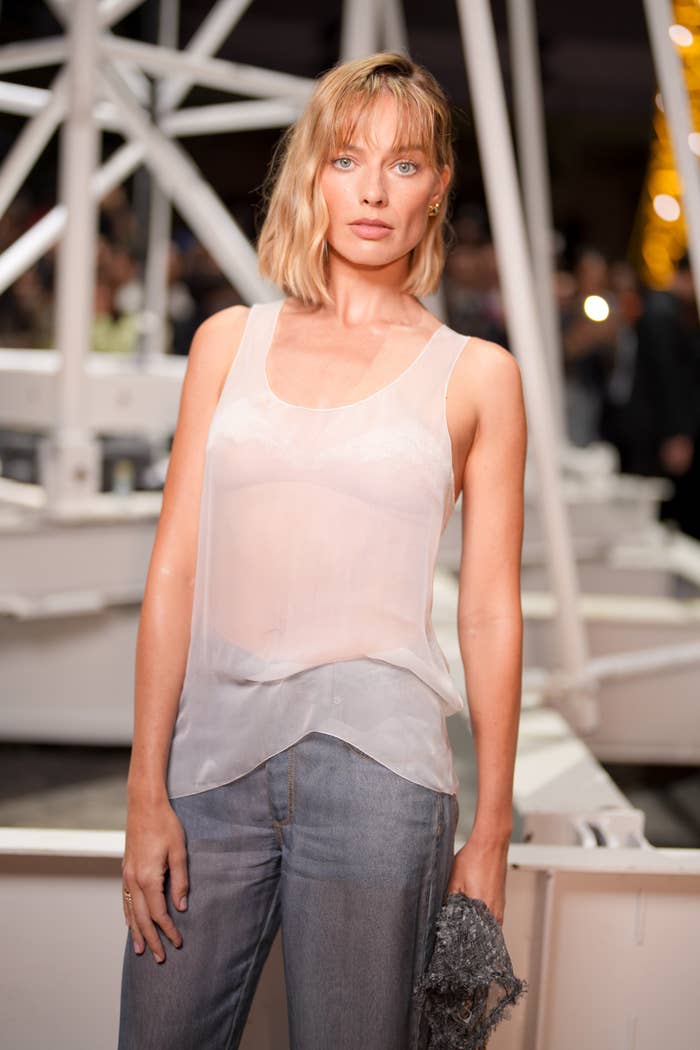 Margot Robbie poses successful  a sleeveless apical  and jeans astatine  an event, lasting  amidst an abstract background, holding a textured accessory