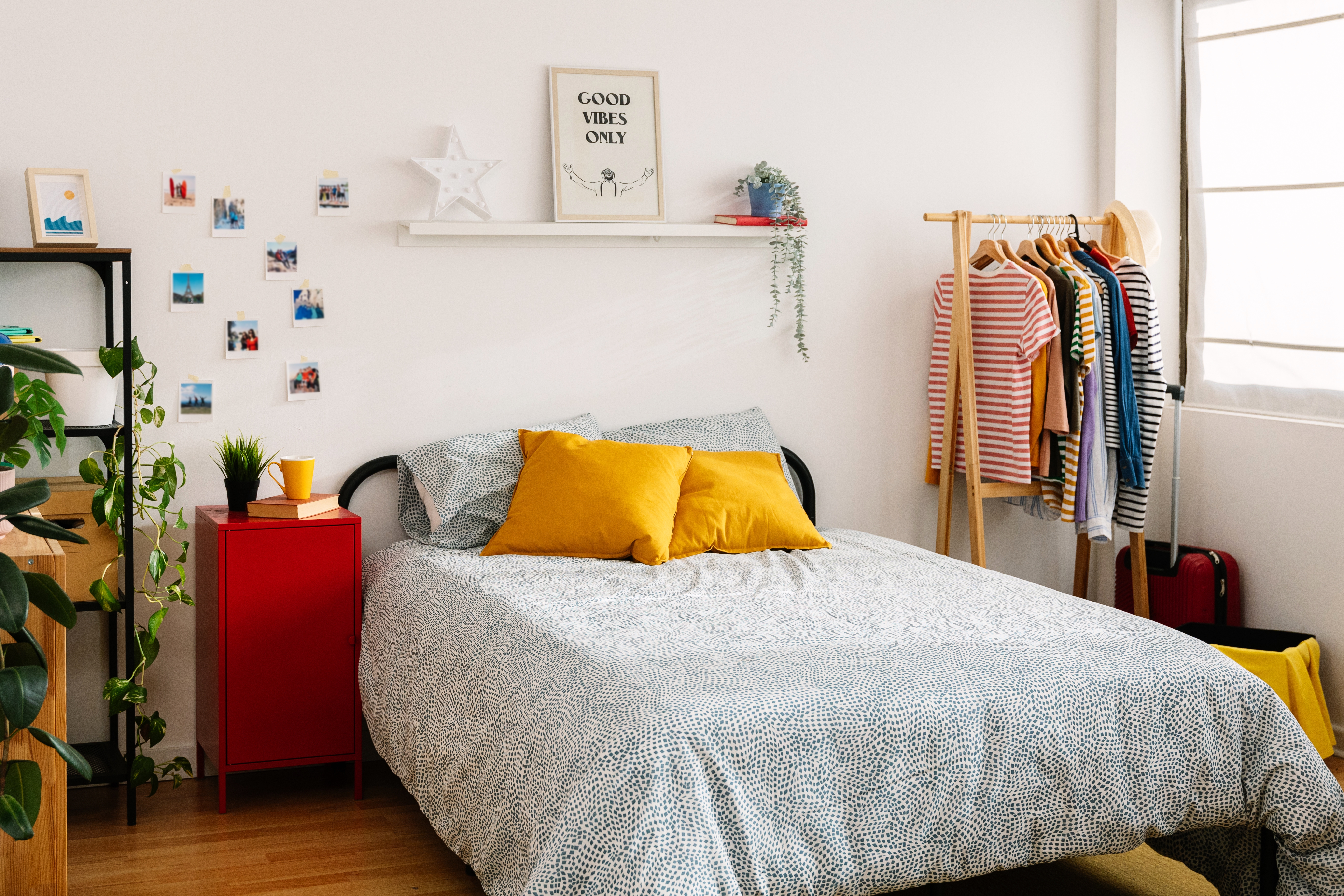 Cozy chamber  with a neatly made bed, pillows, and apparel  connected  a rack, evoking warmth and intimacy. Wall creation  reads "Good Vibes Only."