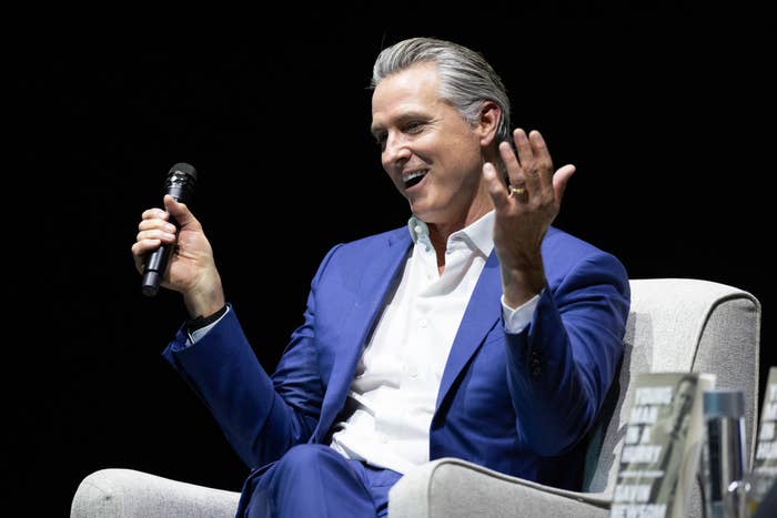 Person successful  a suit   sits connected  a chair, speaking into a microphone, gesturing with 1  manus  during an event