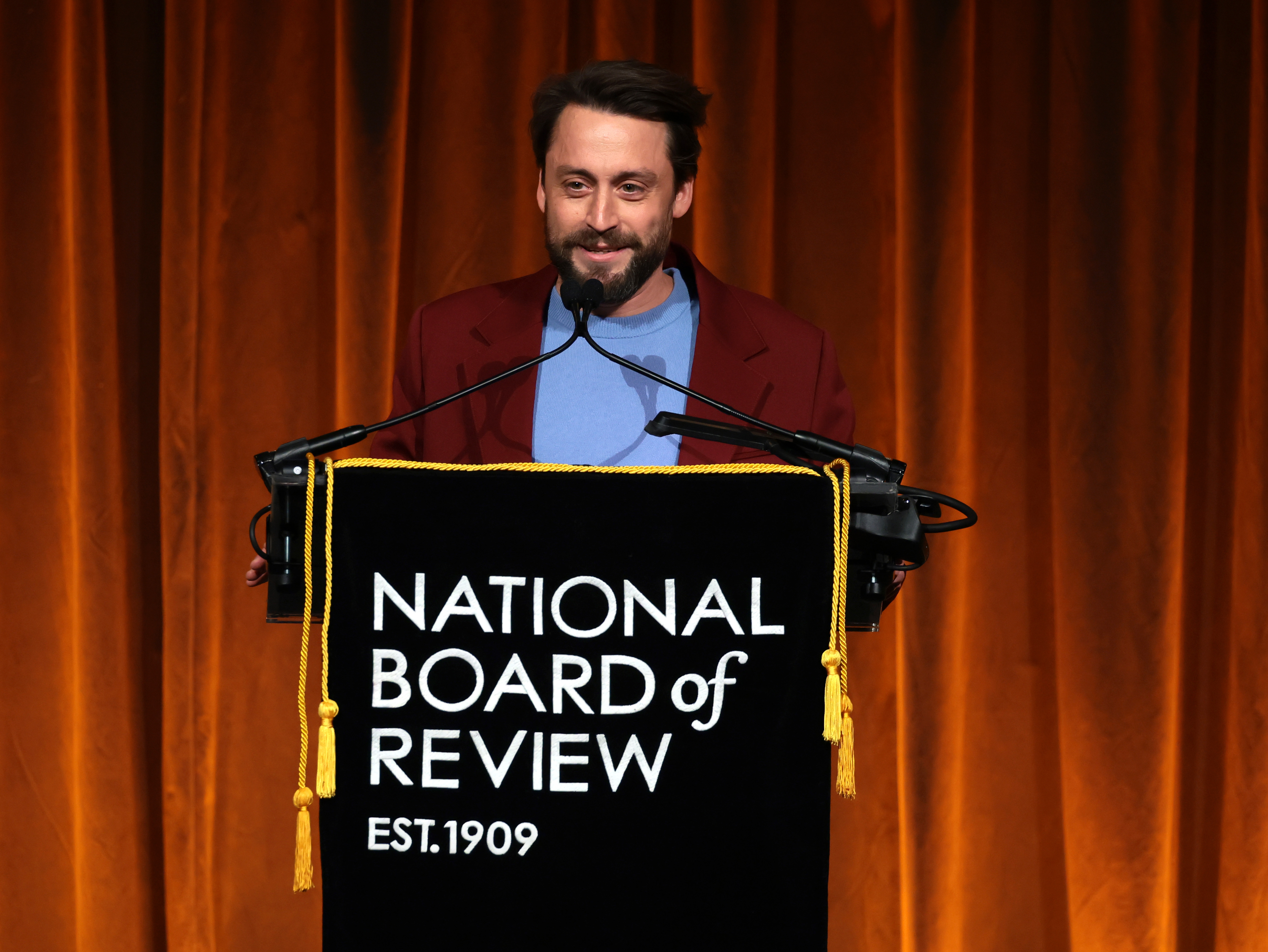 Kieran Culkin stands astatine  a podium with a "National Board of Review" banner, speaking astatine  an event
