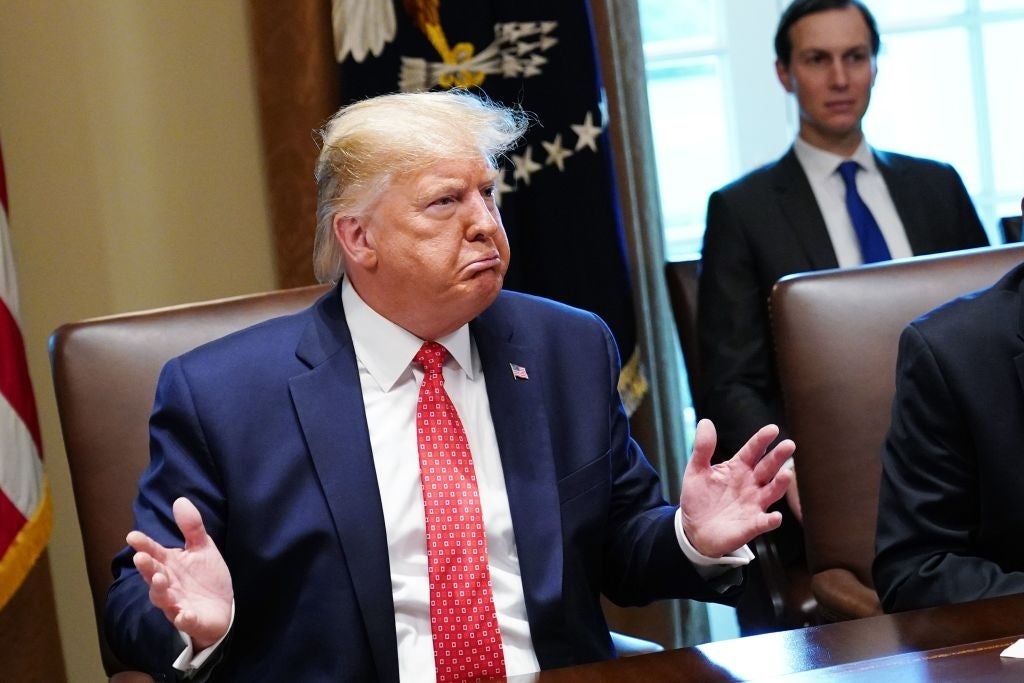 En mann i dress og rødt slips gester mens han sitter ved et konferansebord i en formell setting. En annen person er synlig i bakgrunnen