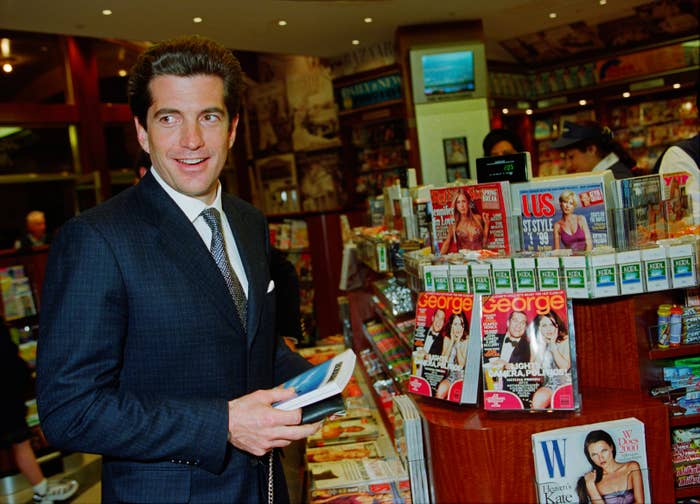 A idiosyncratic   successful  a suit   stands smiling adjacent   a mag  basal   filled with assorted  mag  titles