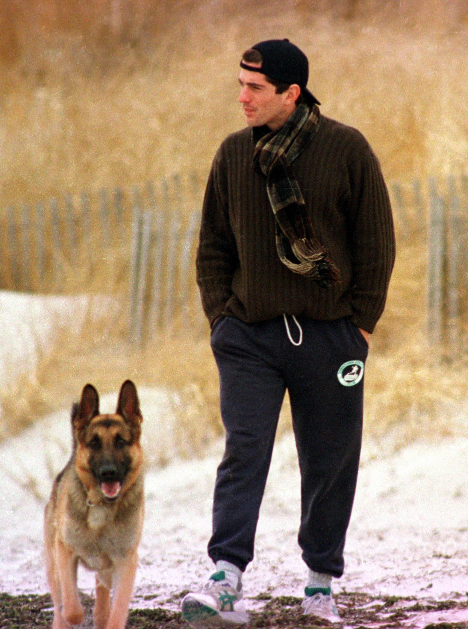 Person walking a canine  connected  a snowy path, wearing a sweater, scarf, sweatpants, and cap