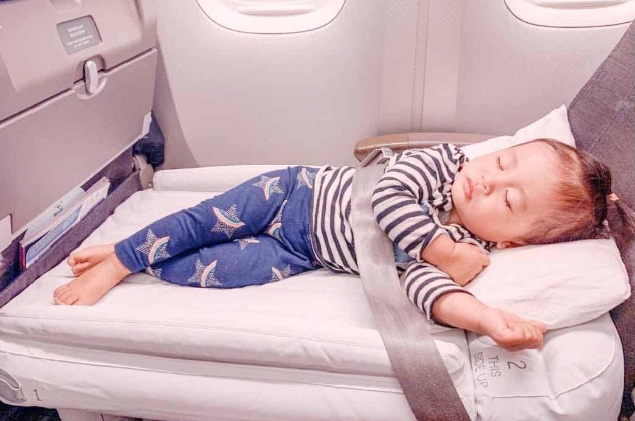 A baby sleeps peacefully on a travel bassinet secured to an airplane seat, wearing a striped top and star-printed pants