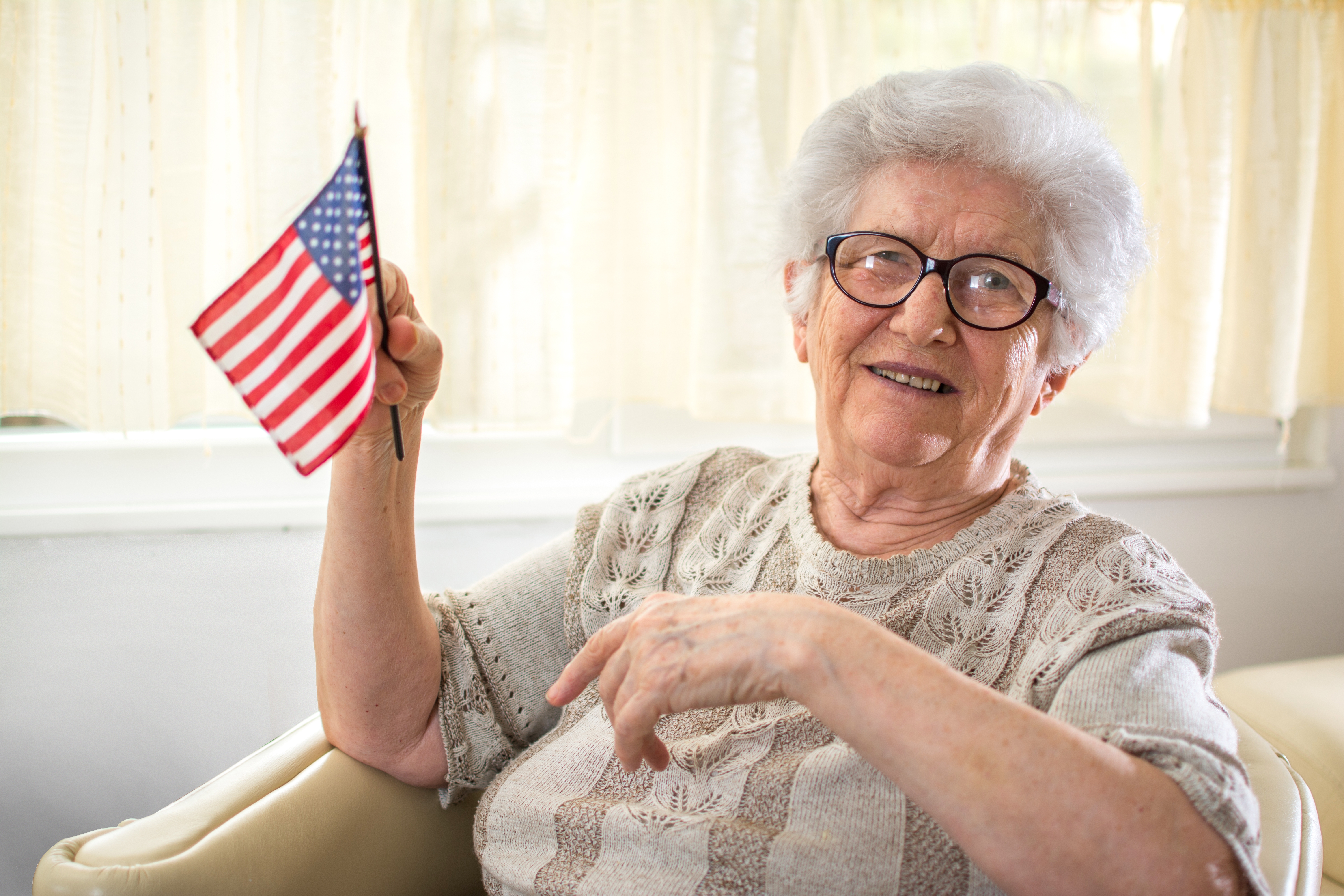 Elderly pistillate   with glasses sitting successful  a chair, smiling, and holding a tiny  American emblem  wrong  a agleam  country   with sheer curtains