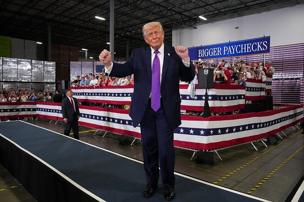 En person i dress gestikulerer selvsikkert på en scene foran et jublende publikum med «Større lønnsslipper»; skilt i bakgrunnen