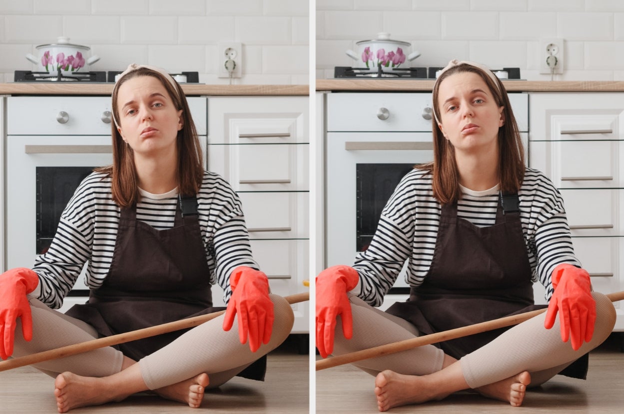Person sitting on kitchen floor, wearing a striped shirt, apron, and red gloves, holding a broom handle. They appear tired or pensive