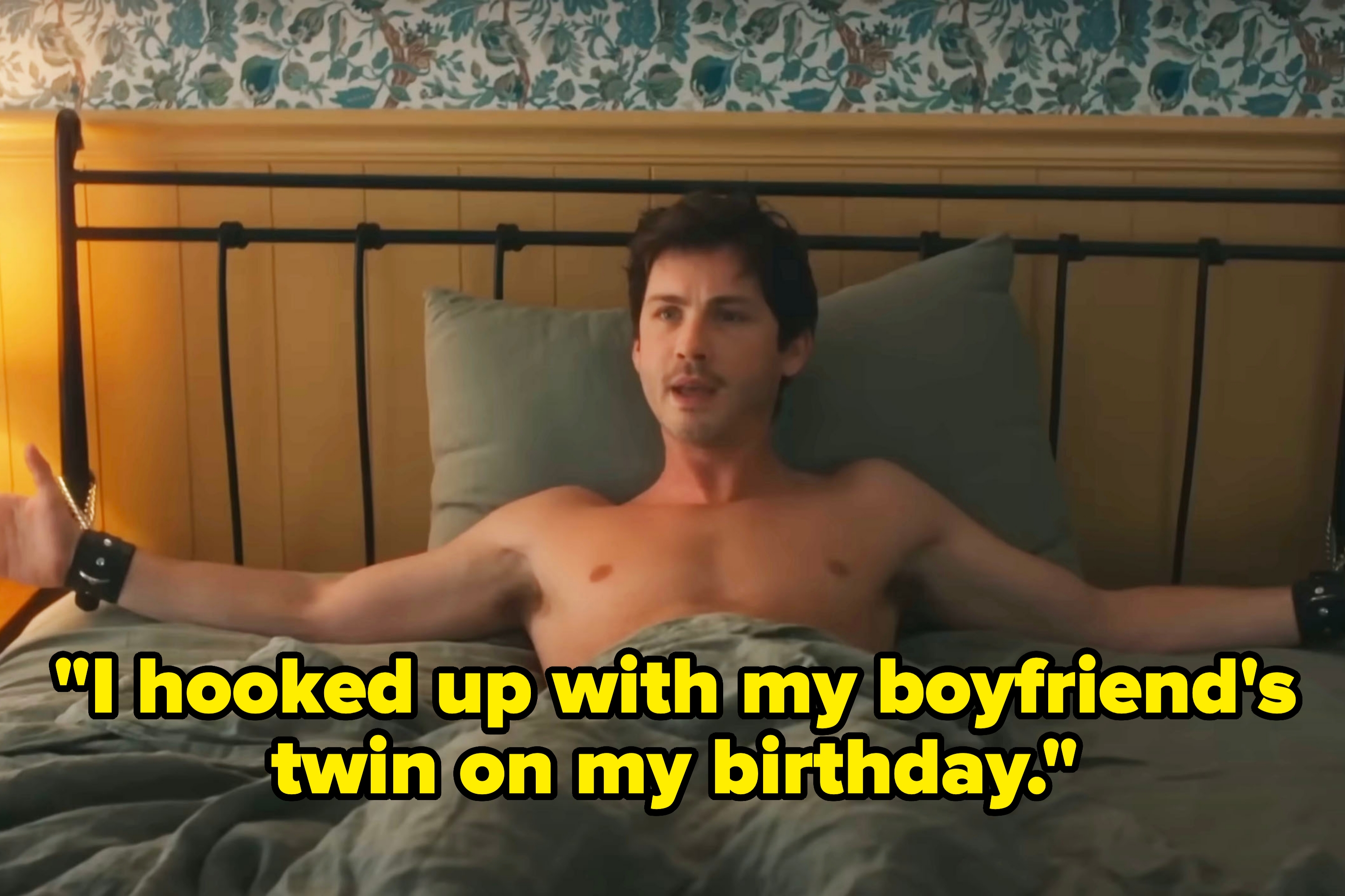 A shirtless man with arms spread wide sits in bed, wrists in cuffs, with a playful expression in a homey bedroom setting