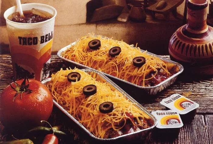 Two Taco Bell Enchirito dishes successful  foil trays, topped with food  and olives, adjacent  to a brushed  drink, tomato, chilies, and taco condiment  packets