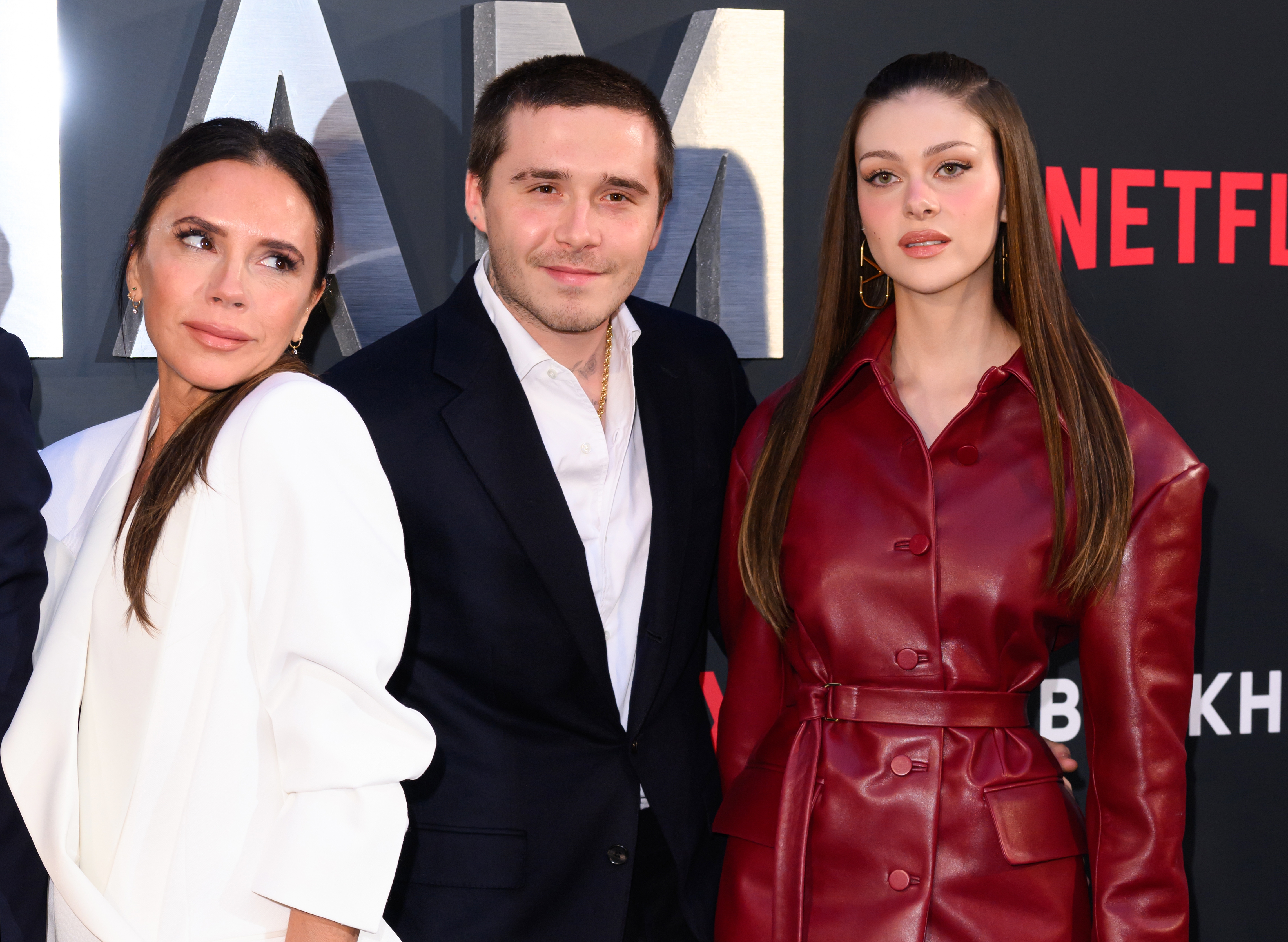 Victoria Beckham successful an elegant blazer and pants stands adjacent to Brooklyn Beckham successful a suit and Nicola Peltz Beckham successful a agelong leather overgarment connected a Netflix reddish carpet