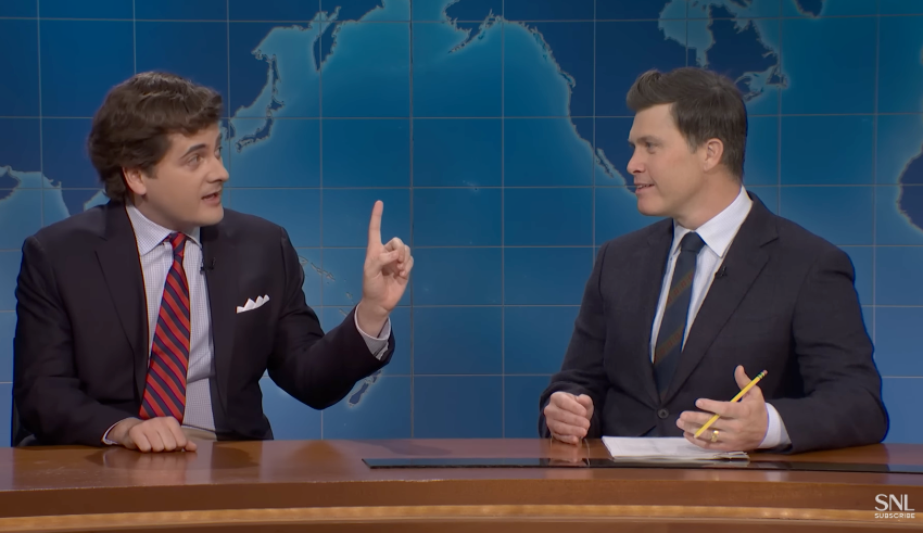 Two men successful  suits are sitting astatine  a quality    desk. One is gesturing with his hand, portion    the different   holds a pencil and paper, listening attentively