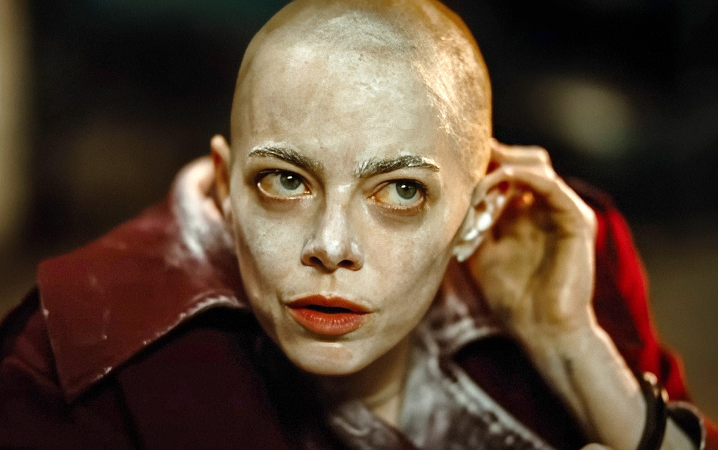 Person med barbert hode, sminke som skaper et dramatisk utseende, justerer øretelefonen. Bruker en formell jakke, virker gjennomtenkt og intens