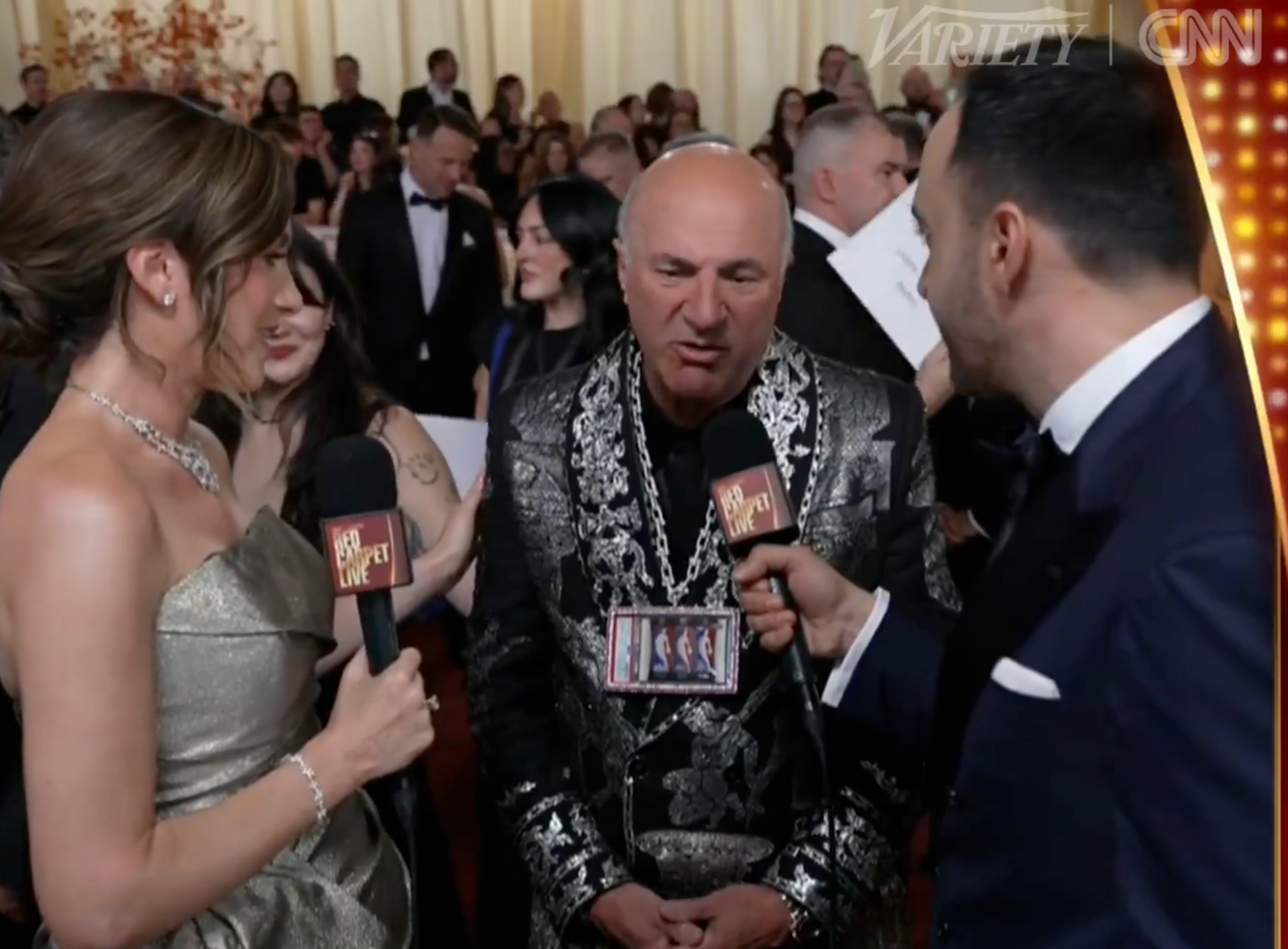 Two reporters interrogation  a antheral   successful  a sequined suit   connected  a reddish  carpet. He wears a necklace with a badge