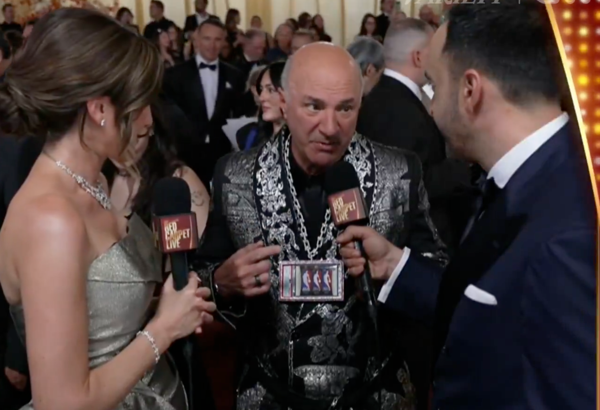 A antheral   successful  a achromatic  patterned suit   with a ample  concatenation  accessory is being interviewed connected  a reddish  carpet by 2  reporters holding microphones