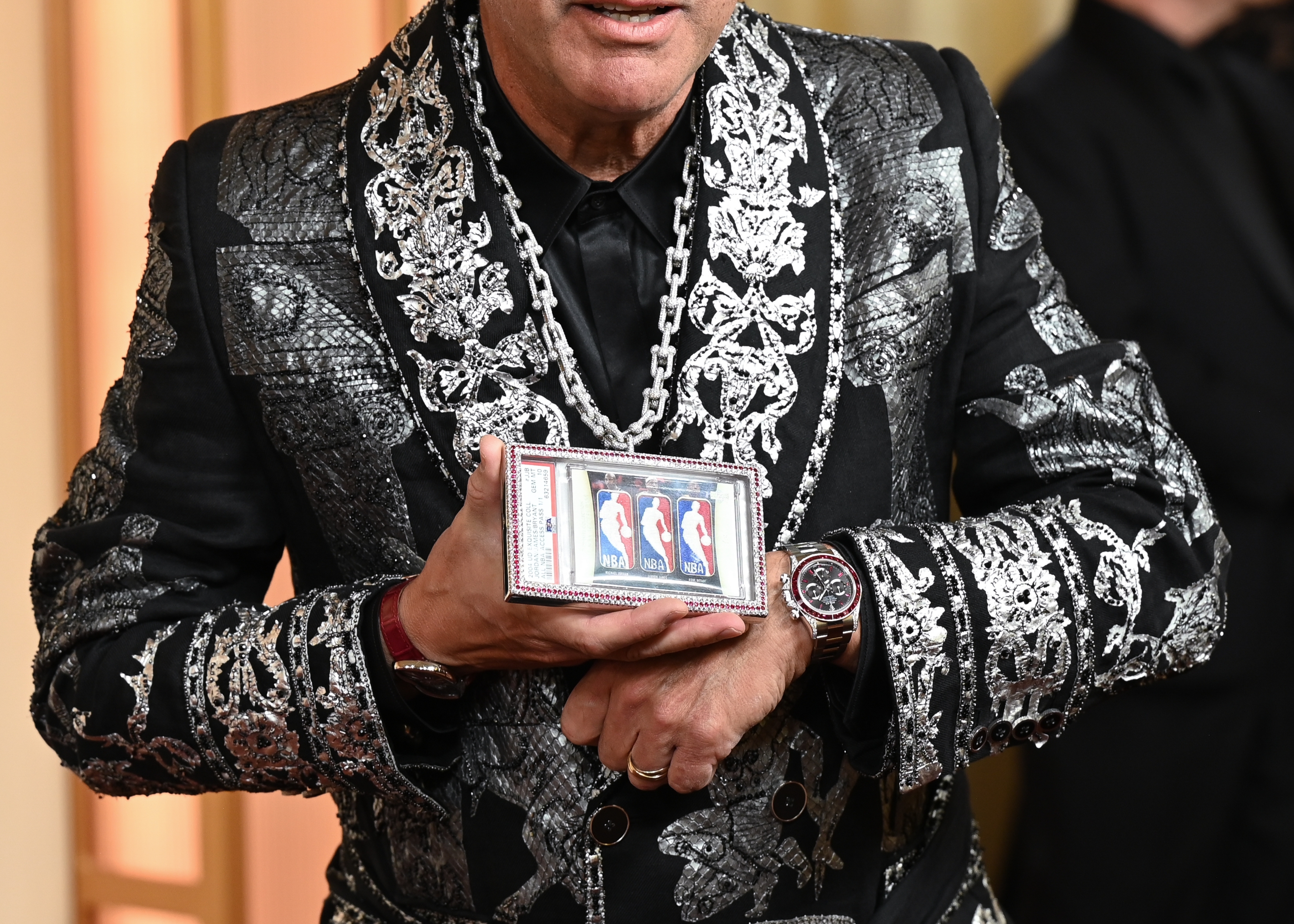 Man connected  reddish  carpet wearing an ornate, patterned suit, holding a telephone  displaying a recognition  paper  image. People and a camera are successful  the background