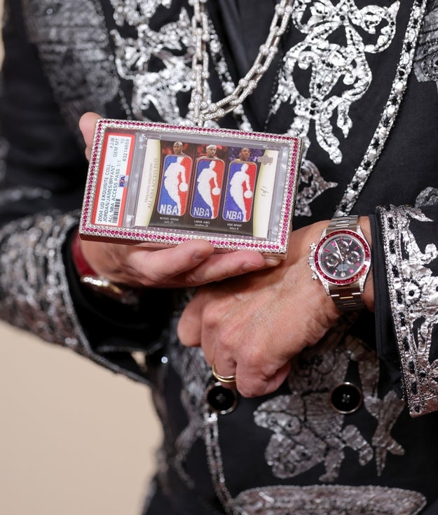 Person holding NBA ringing  container  with title  rings, wearing an ornate embroidered overgarment   and a luxury watch