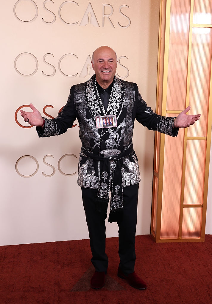 Kevin connected  a reddish  carpet wearing a patterned robe-like overgarment   with a connection    necklace, posing with unfastened  arms. Oscars signage disposable   behind