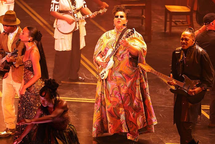 Performers connected  stage, 1  successful  a vibrant patterned robe holding a guitar, others with instruments and dancing, creating a lively philharmonic  scene