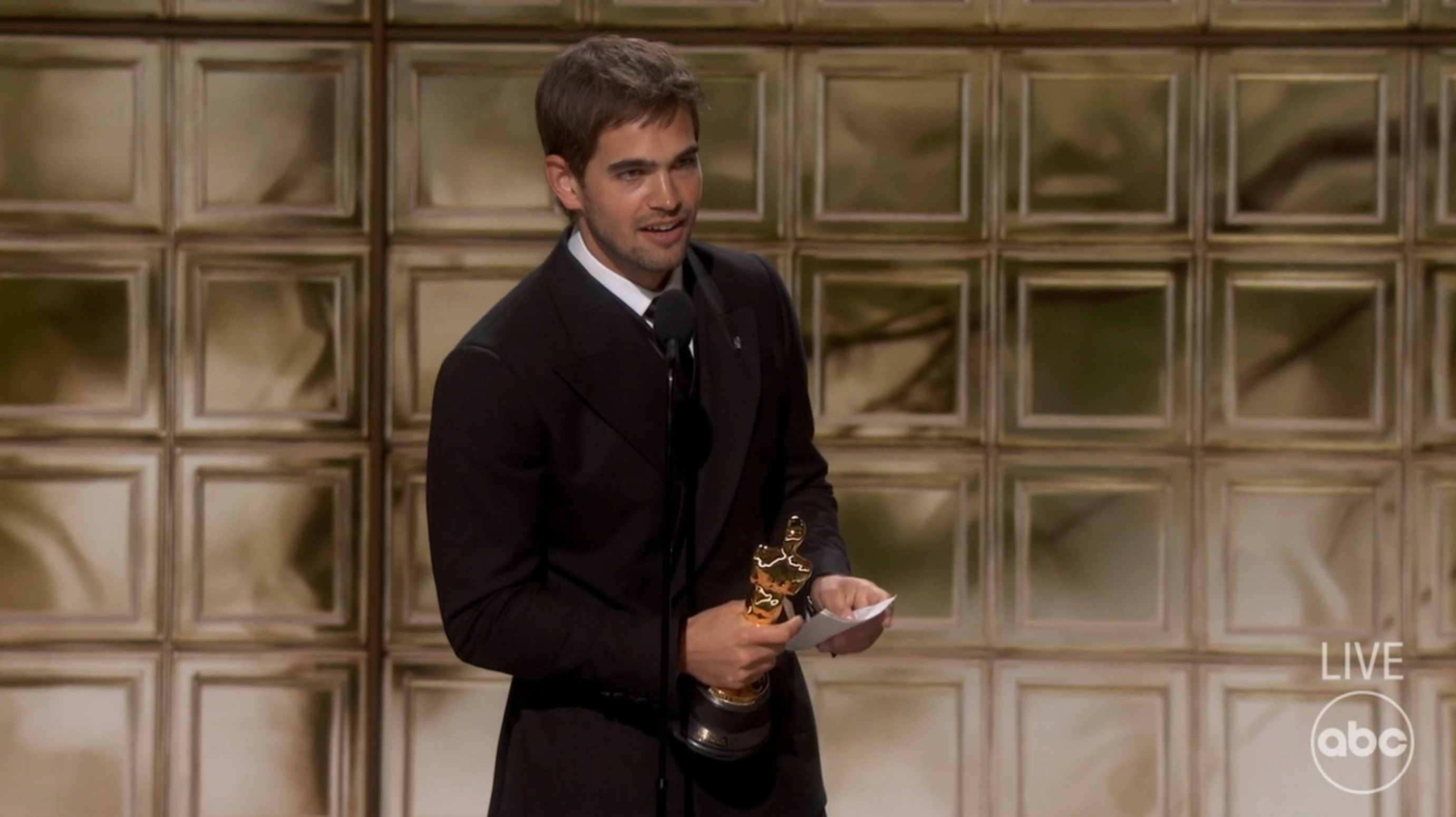 A antheral successful a acheronian suit holds an grant statue portion speaking connected signifier astatine a televised event. The inheritance features a geometric pattern