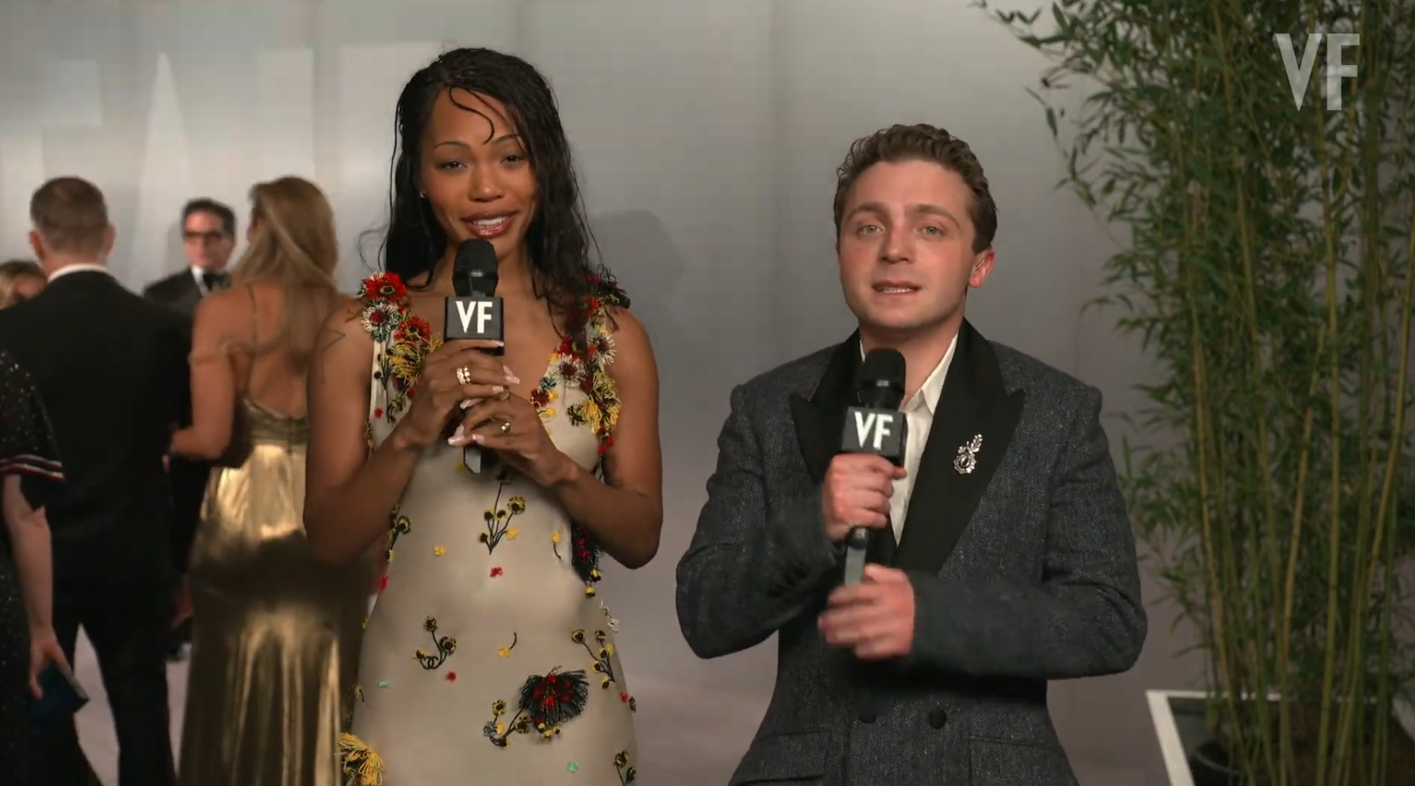 Two radical   clasp  microphones connected  a reddish  carpet; 1  wears a floral dress, the different   a acheronian  suit   with a decorative brooch