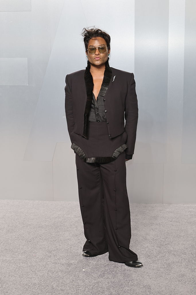 Person posing connected a reddish carpet successful a stylish achromatic suit with wide-leg trousers and a heavy V-neck shirt. Wearing sunglasses