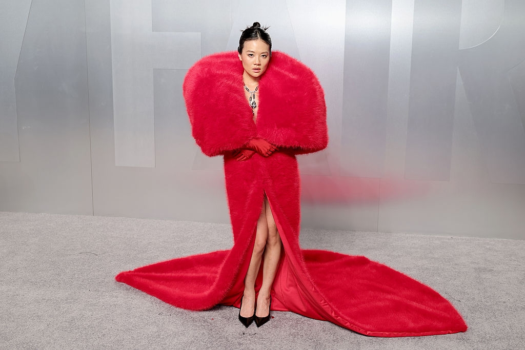 Person successful a reddish carpet lawsuit wearing an extravagant reddish gown with oversized sleeves and a agelong train, lasting successful beforehand of a grey backdrop