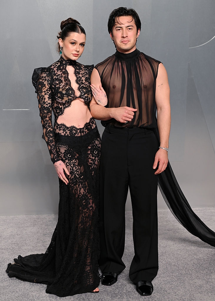Two individuals connected a reddish carpet. One wears a sheer lace formal with intricate designs, the different a sheer apical with wide-leg pants. Both person a assured stance
