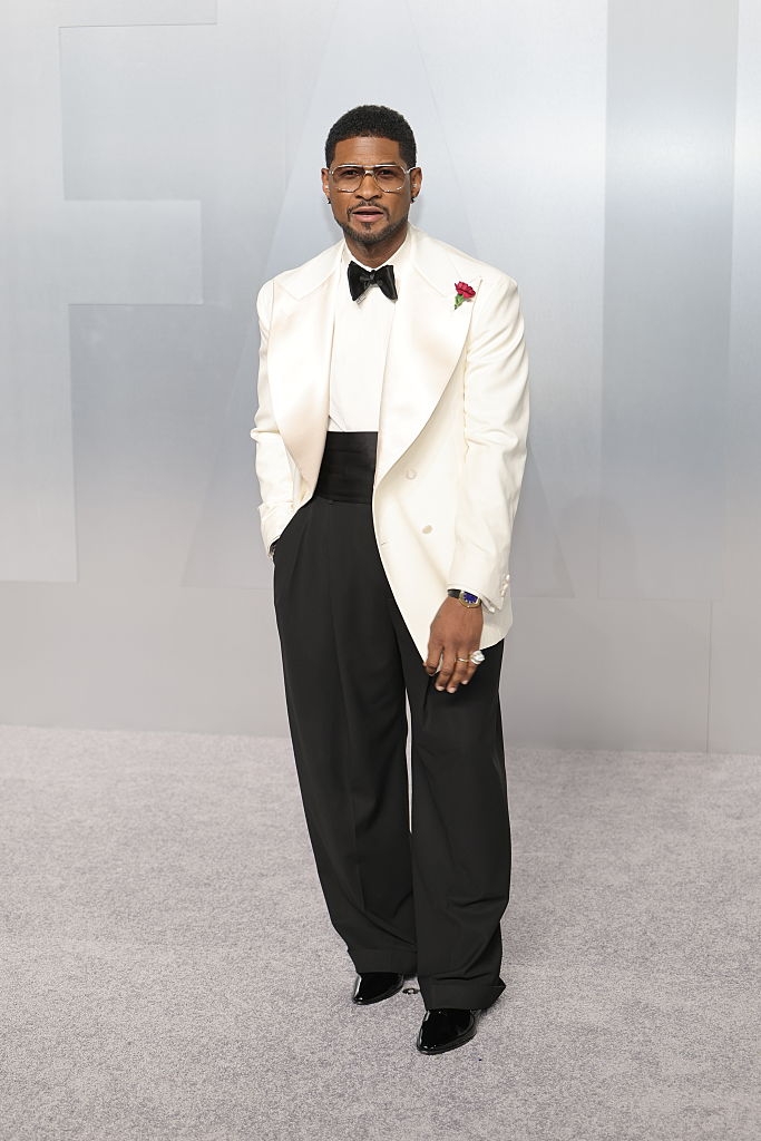 Person wearing a ceremonial achromatic blazer, achromatic bow tie, and trousers, with a reddish floral lapel pin, stands connected a grey carpet