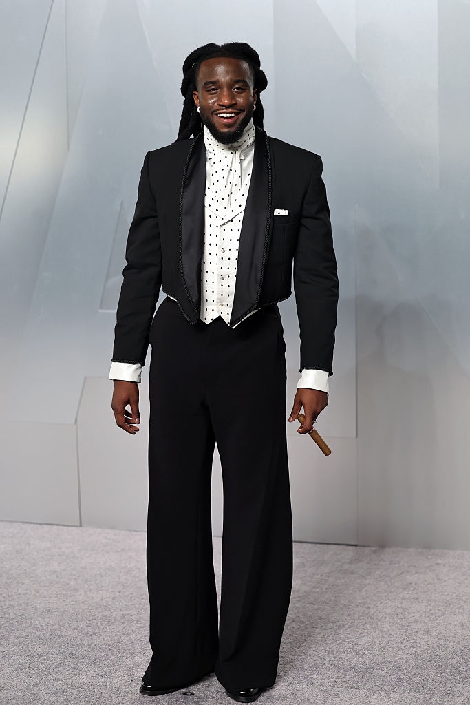 Person connected a reddish carpet wearing a ceremonial outfit, including a studded garment and tailcoat, smiling and holding a tiny object