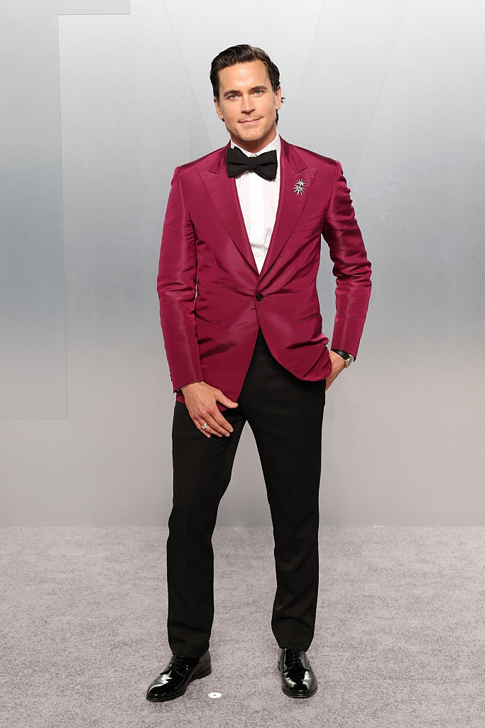 Person connected reddish carpet wearing a crisp suit with a burgundy jacket, achromatic bow tie, and achromatic pants, posing confidently