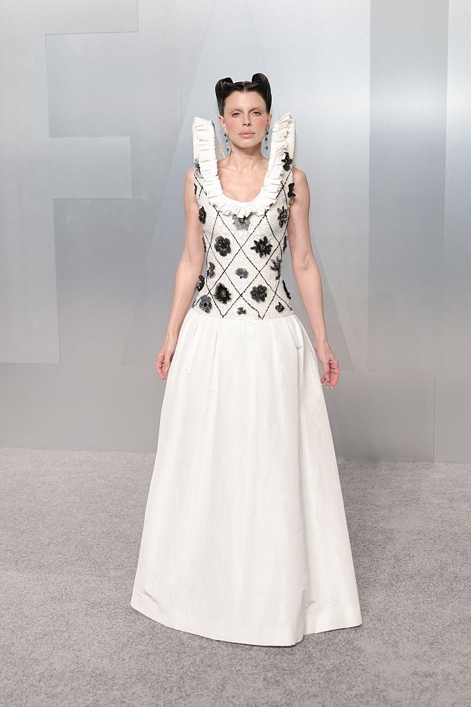 Person successful a floor-length, sleeveless gown with floral patterns and a ruffled neckline, posing connected an lawsuit carpet