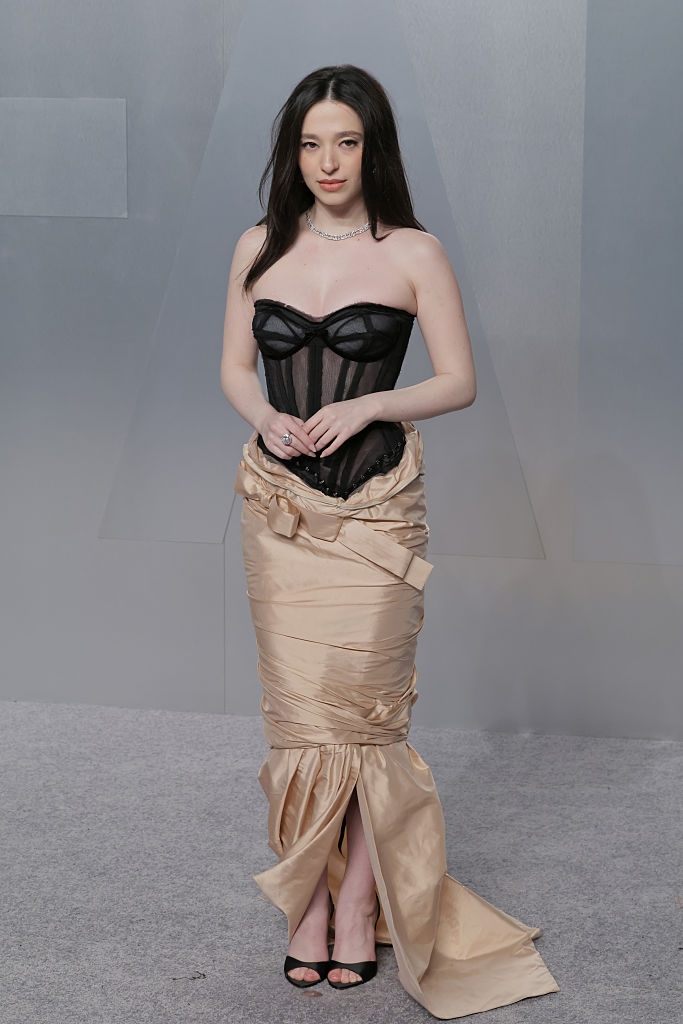 A idiosyncratic poses connected a reddish carpet wearing a structured achromatic corset apical with a satin, draped floor-length skirt