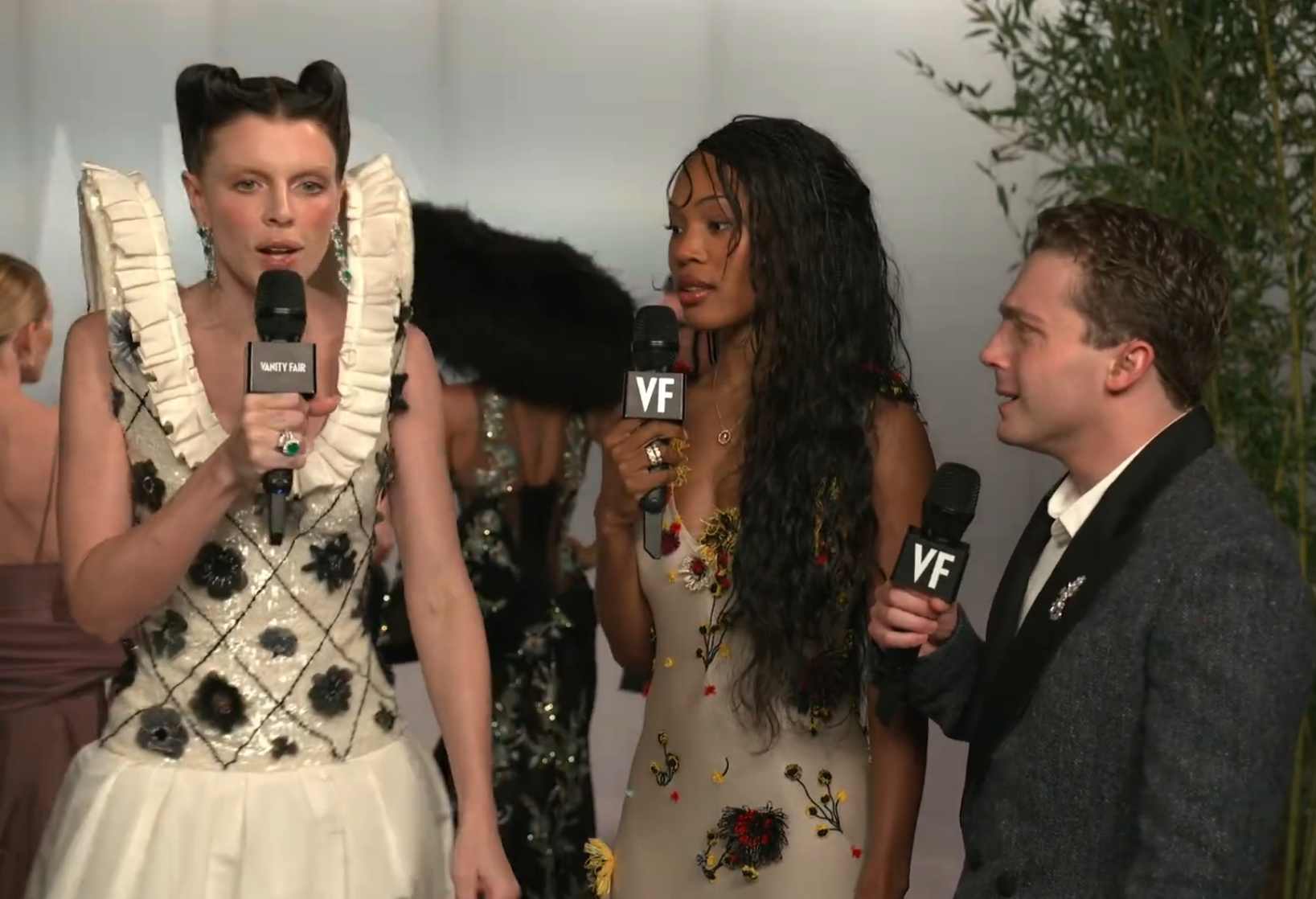 Three radical holding microphones with "VF" logos astatine an event. One wears a ruffled dress, different a floral gown, and the 3rd a suit